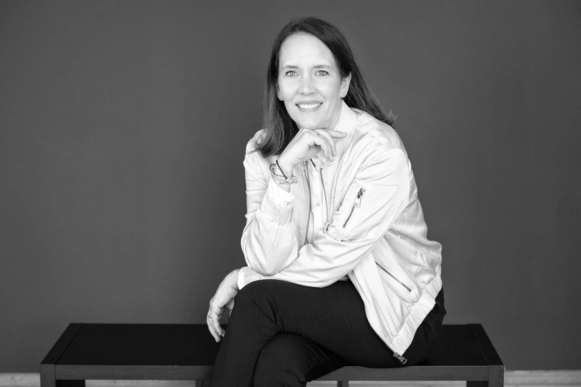Schwarz-weiße Portraitaufnahme einer lächelnden Frau mit dunklem Haar, sitzend auf einer schwarzen Bank, vor einem dunklen Hintergrund, trägt ein zartes top und eine Jacke, ihre Hand ruht unterhalb des Kinns.