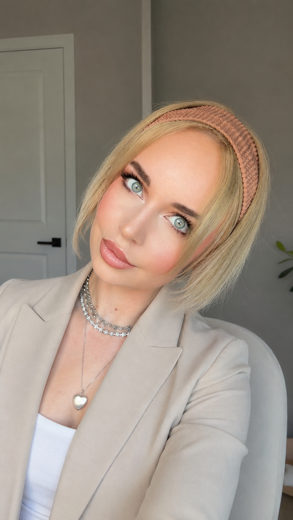 A woman with blonde hair, blue eyes, and wearing a pink headband, beige blazer, white top, layered necklaces, and soft makeup, sitting in a room with light-colored walls and a white door.