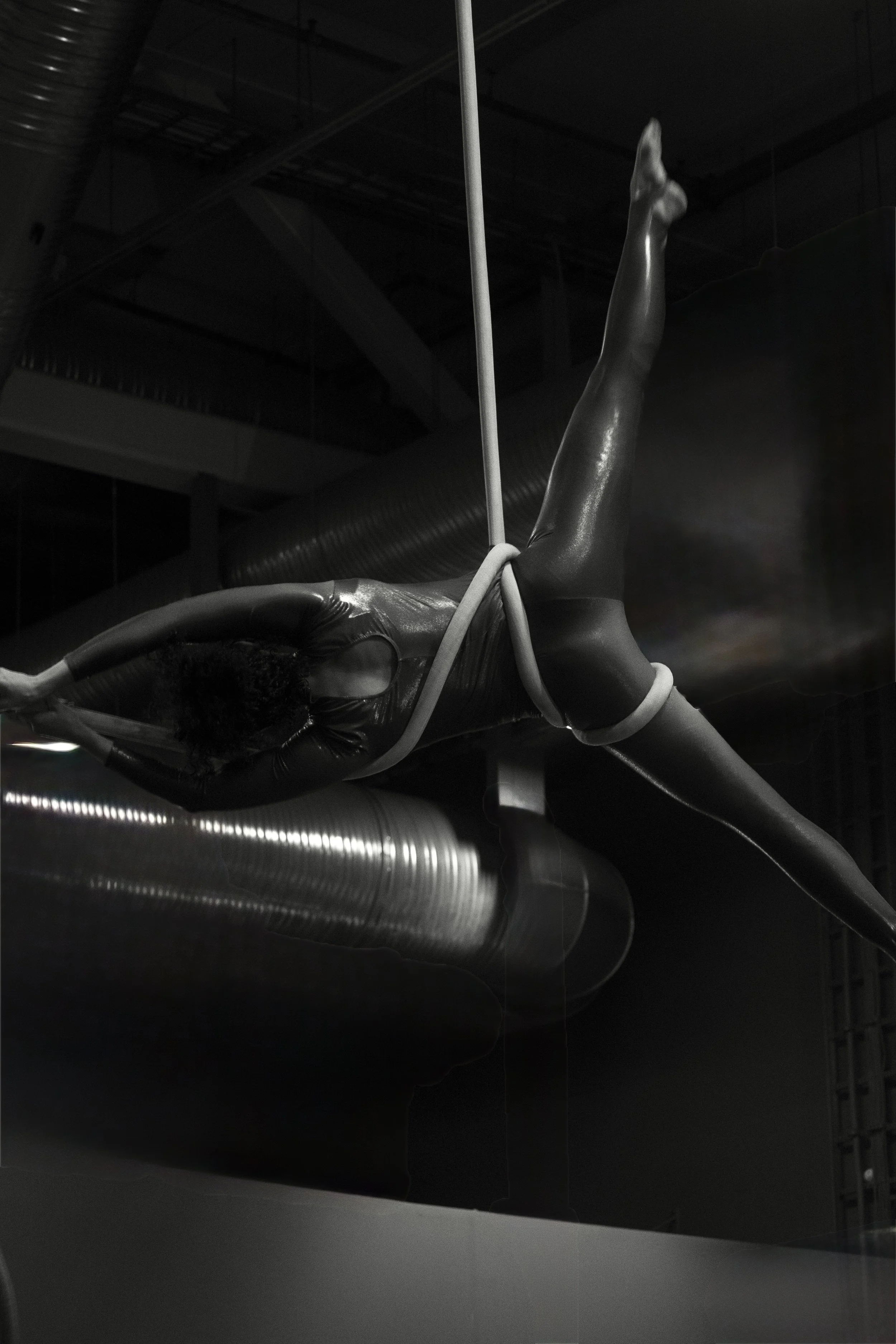 A shiny, metallic gymnast in black clothing performing an aerial routine, holding onto a white rope with both hands and extending one leg upward, in a dark industrial-style gymnasium.