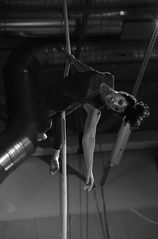 A woman performing an aerial rope dance move on a vertical rope in a studio with industrial ceiling pipes and lighting.