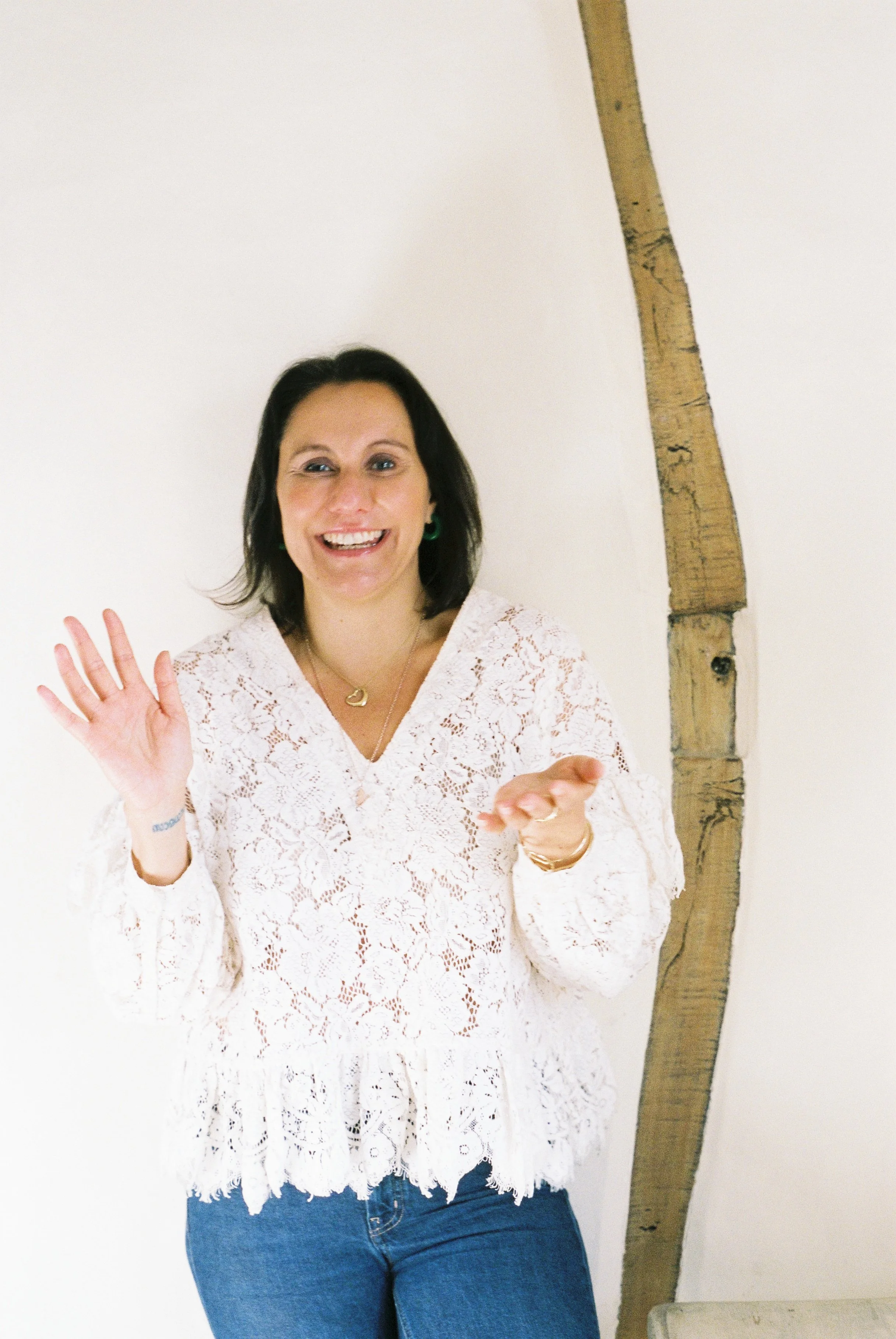 A smiling woman with dark hair wearing a white lace top and blue jeans, waving with her right hand, standing against a white wall with a wooden beam on her right side.