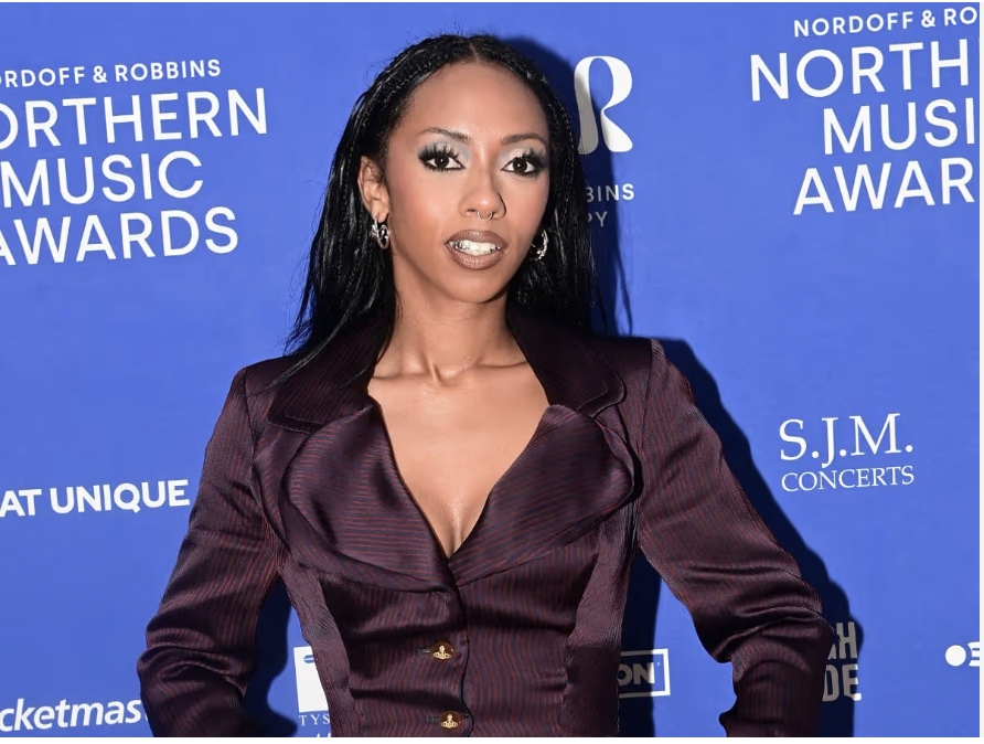 A woman with long black hair and earrings posing at a music awards event, standing in front of a blue backdrop with white text.