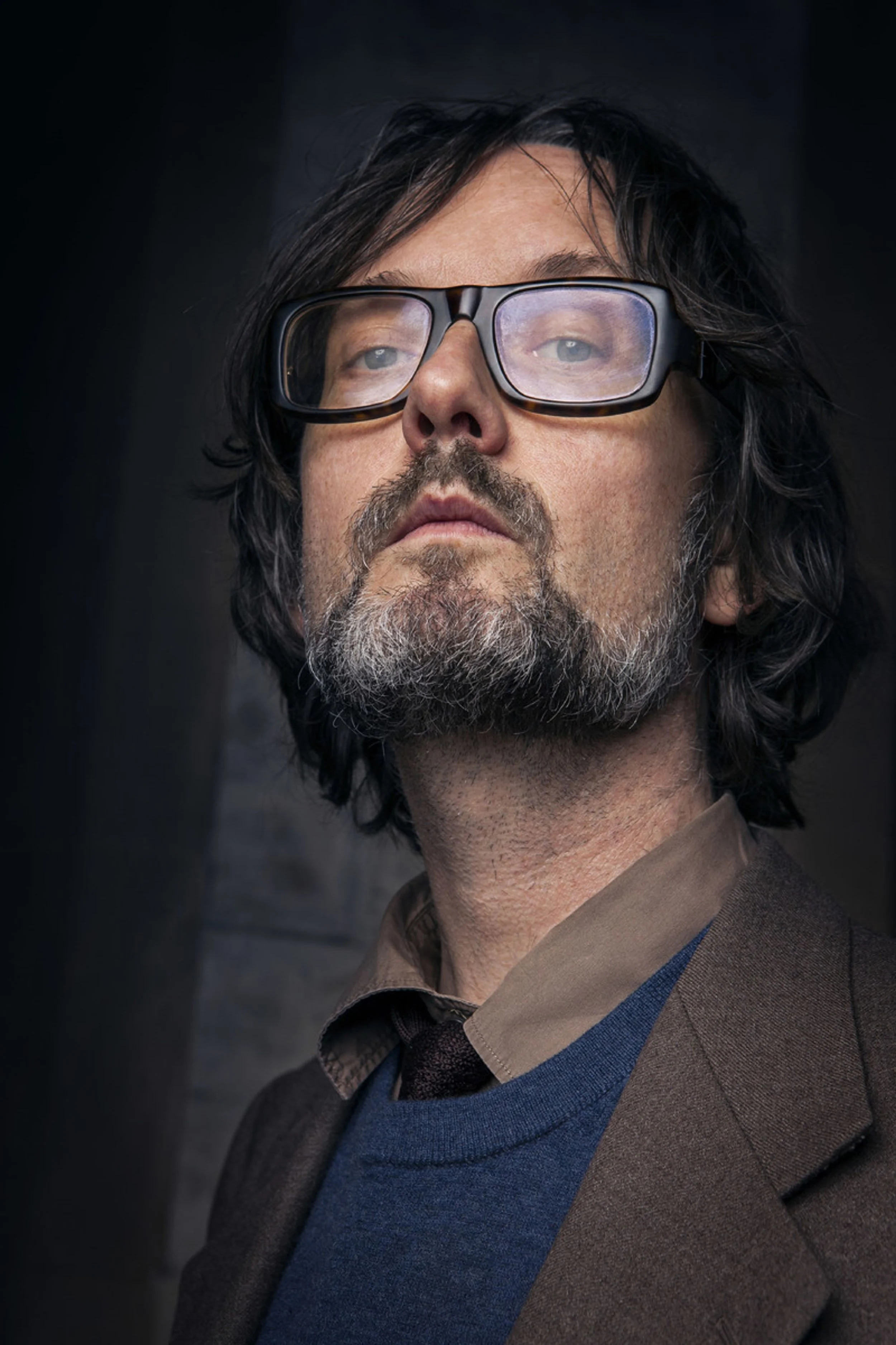 Close-up portrait of a man with glasses, dark hair, and a beard, wearing a brown coat with a blue sweater and a darker shirt underneath, against a dark background.