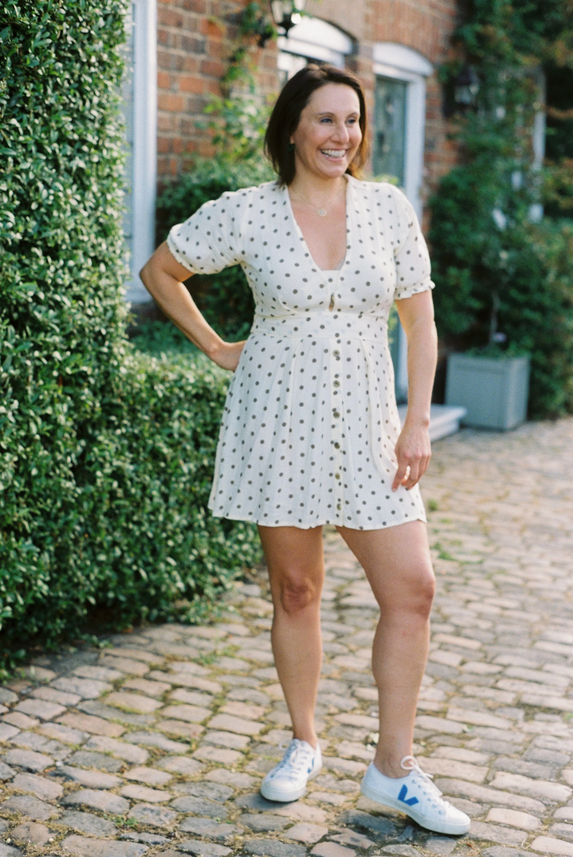 A woman standing on a cobblestone pathway outdoors, smiling, in a white dress with black polka dots and white sneakers, with greenery and brick buildings in the background.