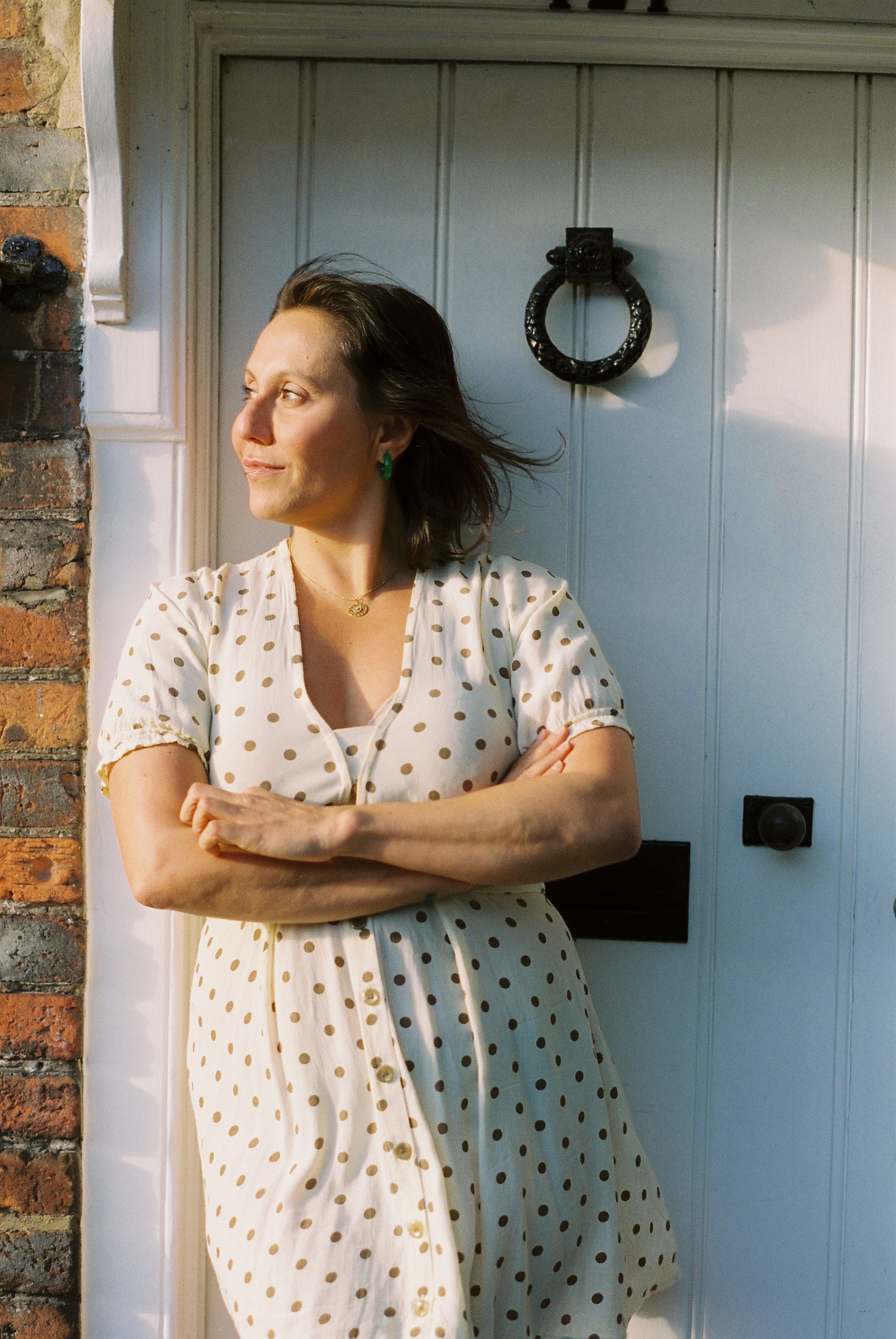 A woman with shoulder-length dark hair wearing a white vintage style dress with brown polka dots, standing with crossed arms outside near a wall and door, looking to her left, with sunlight illuminating her face.