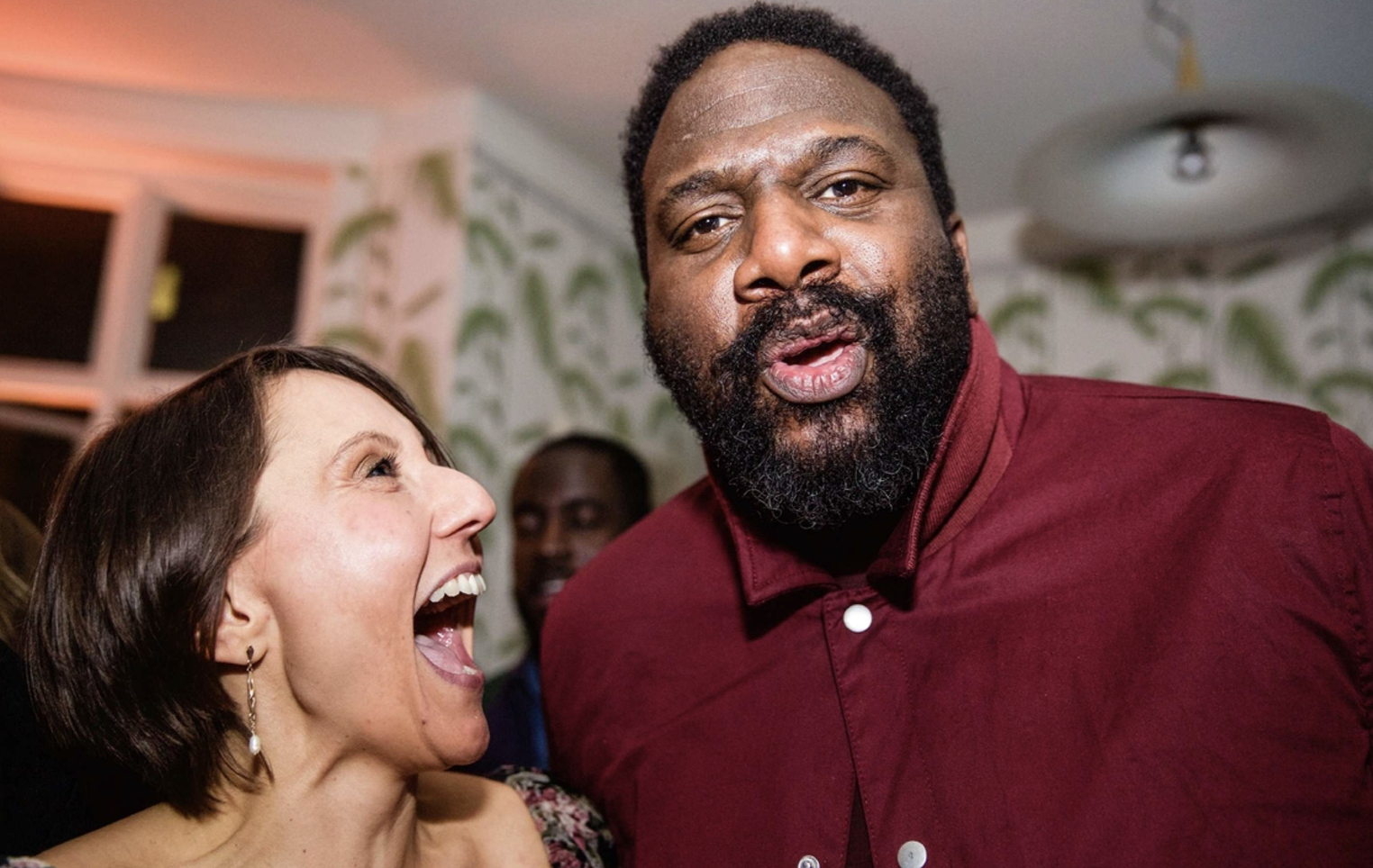 A woman with short brown hair laughing and a man with a beard and short curly hair making a surprised facial expression at a social gathering inside a room with a patterned wall.