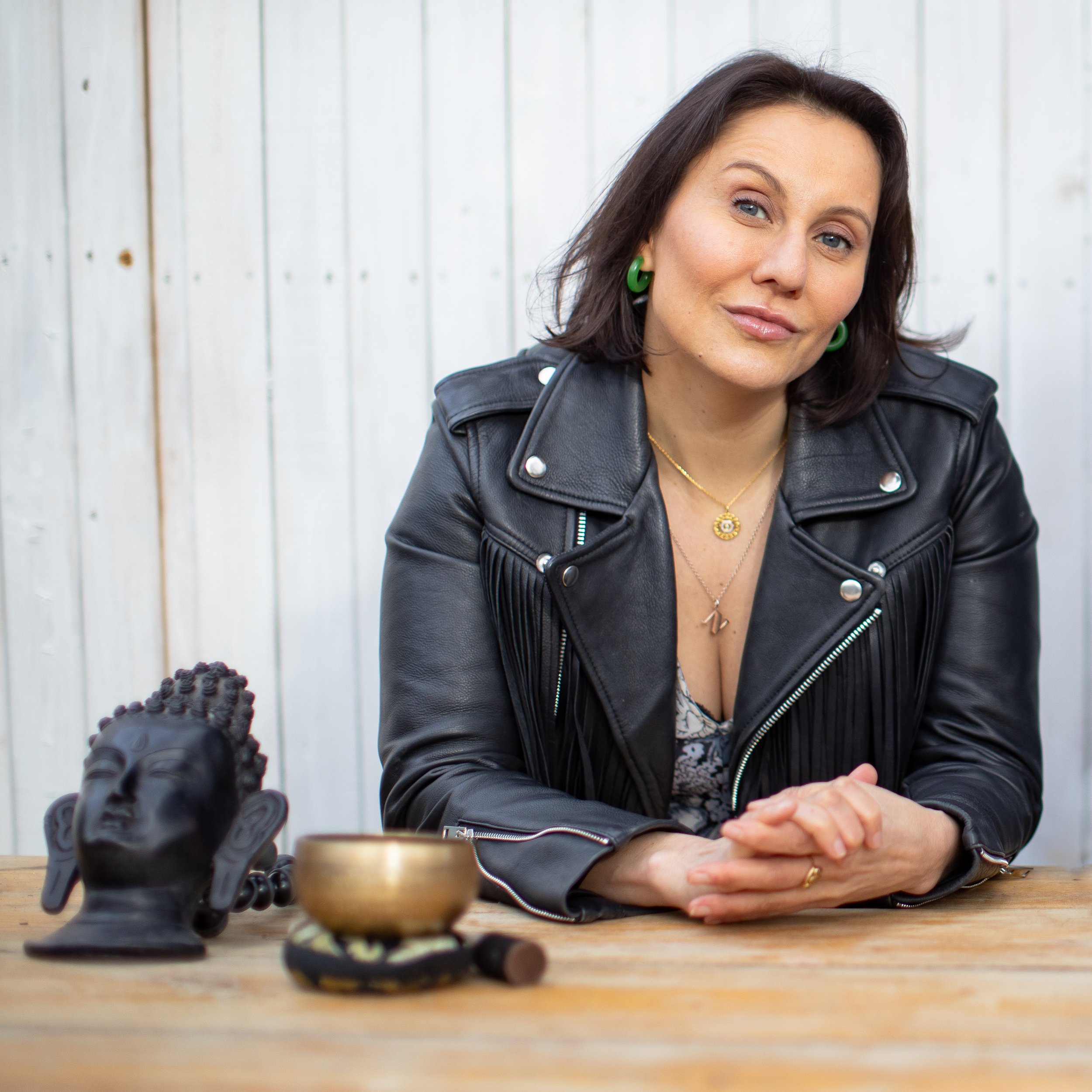 A woman with dark hair and blue eyes wearing green earrings and a black leather jacket, sitting at a wooden table with a chess set in the foreground