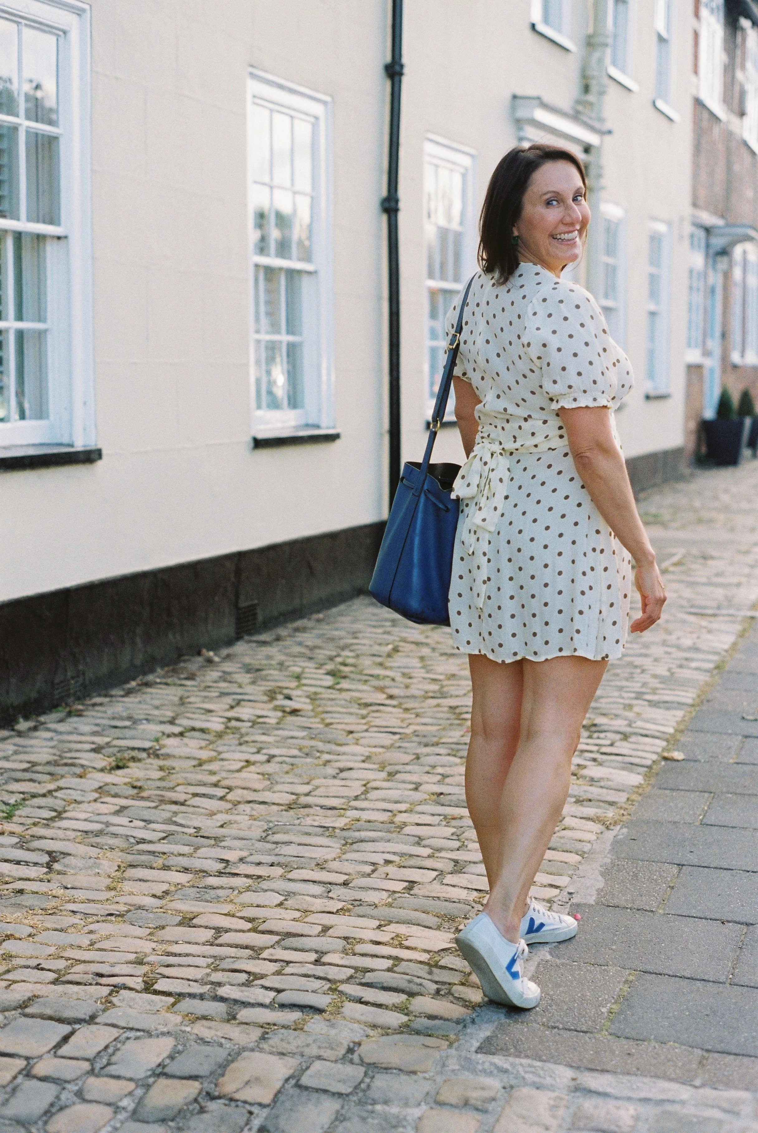 A woman smiling and walking on a cobblestone street, wearing a white polka dot dress and sneakers, heading away from the camera, with a row of houses lining the street.