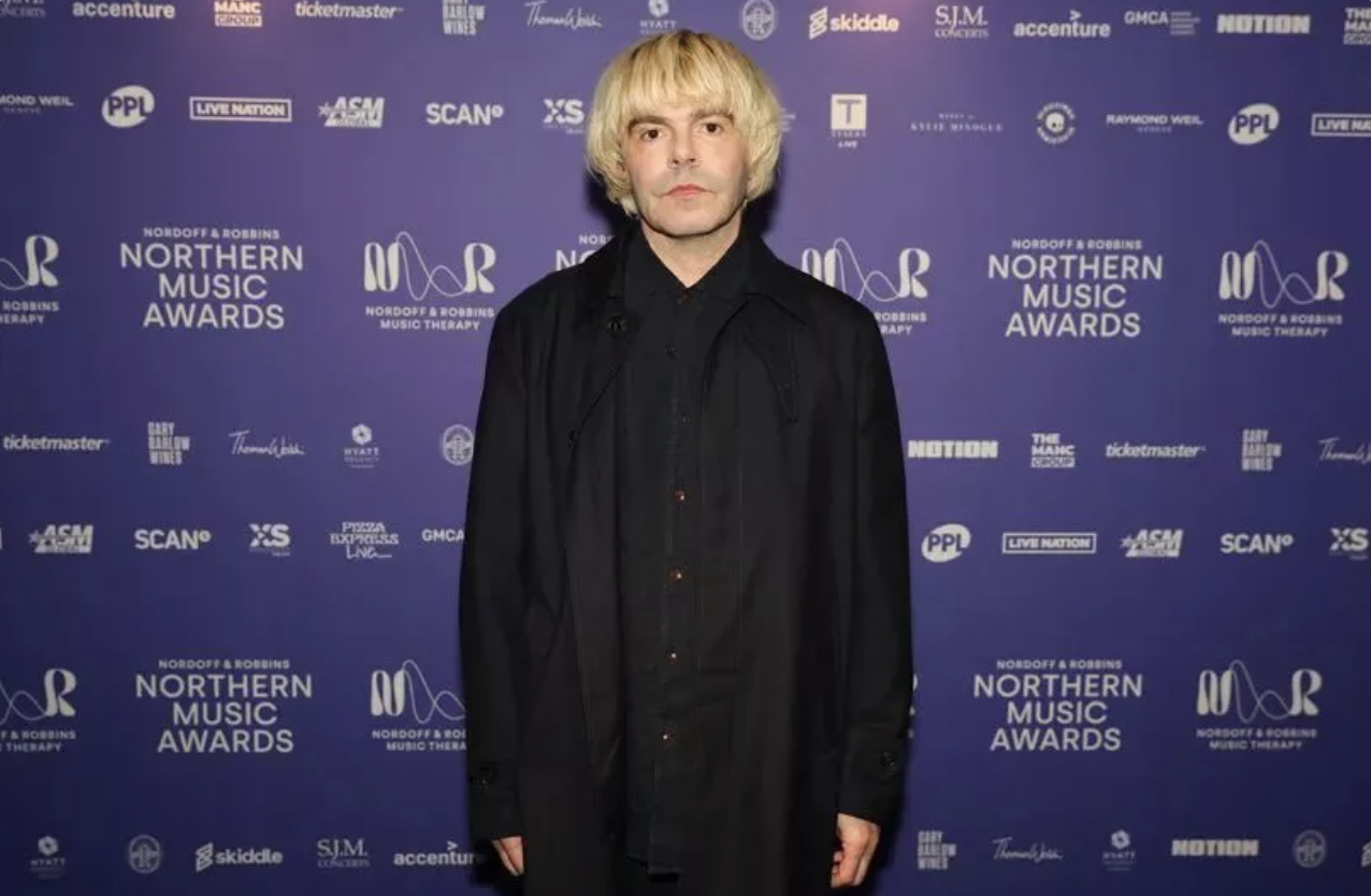 A person with blonde hair wearing a black coat standing in front of a purple backdrop with event logos and text for the Northern Music Awards.