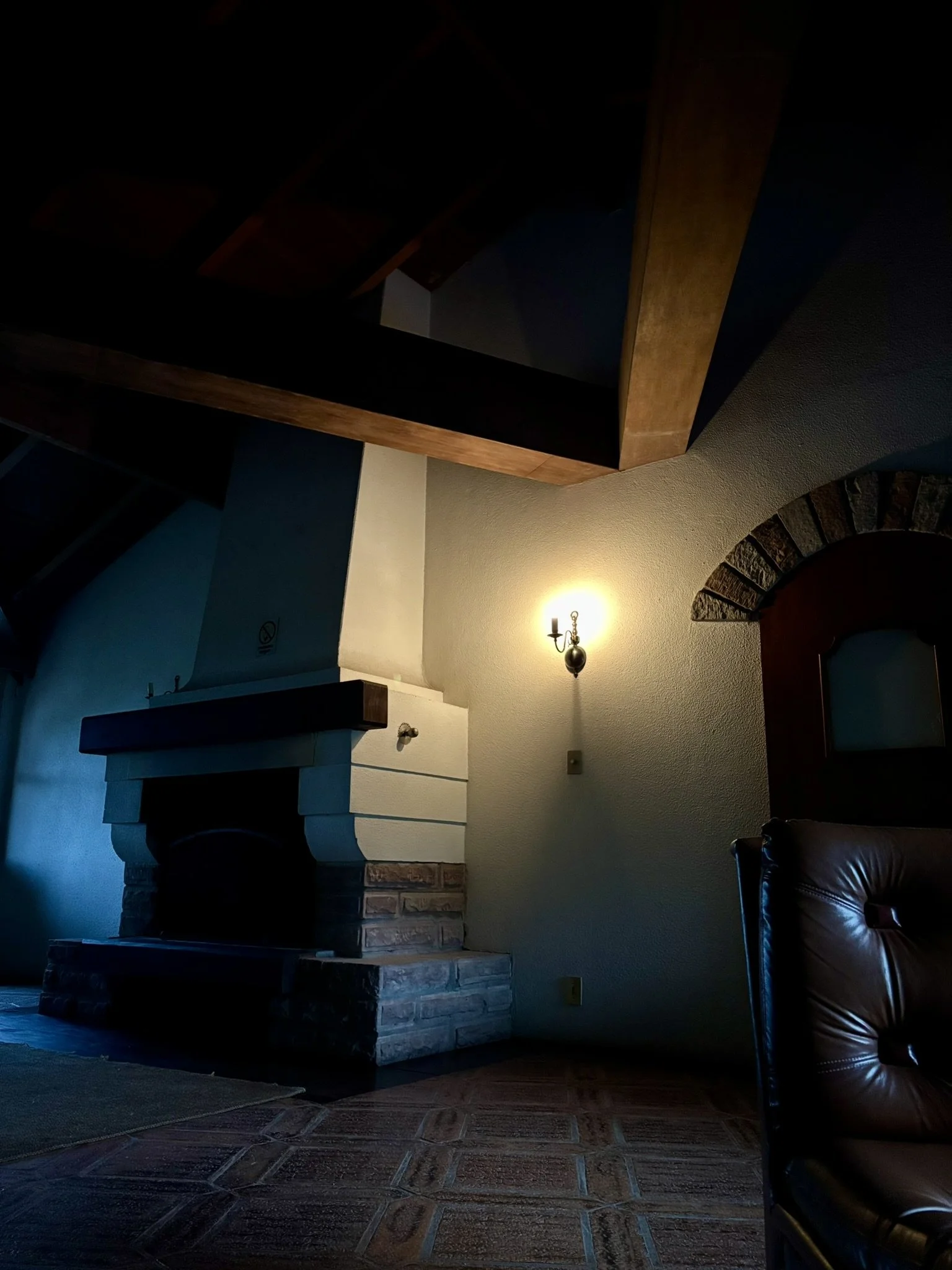 Sala de estar com lareira de pedra, parede amarela, luminária antiga, piso de ladrilhos, cadeira de couro marrom, teto de madeira com vigas aparentes e arco de pedra na porta ao fundo.