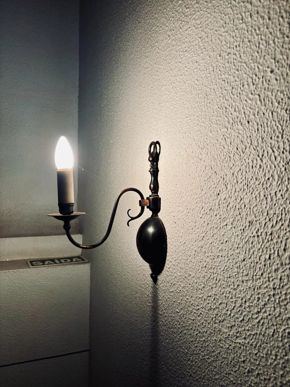 Luminária de parede antiga com vela acesa, fixada na parede com textura áspera.