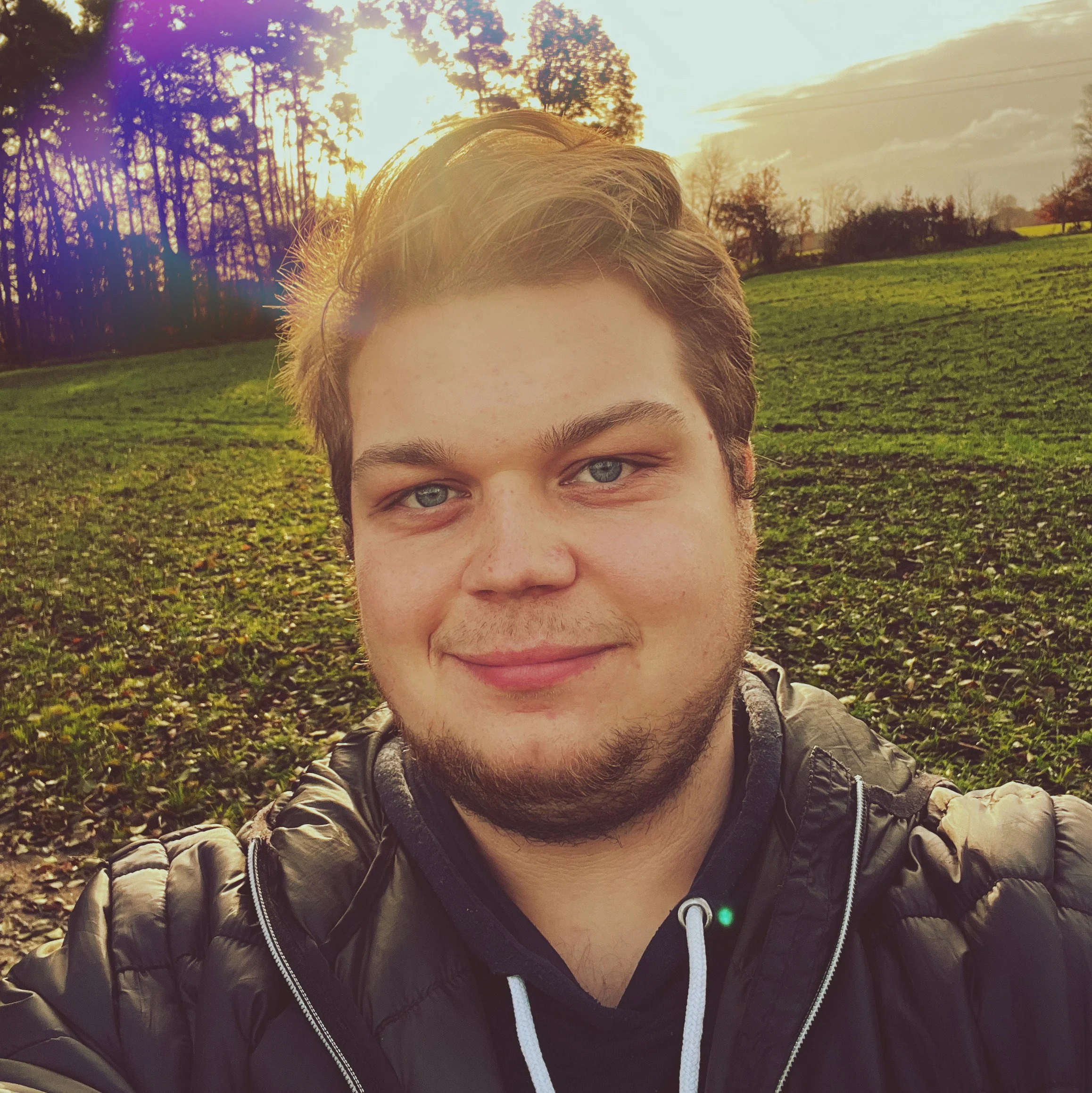 Nahaufnahme eines jungen Mannes mit blauen Augen und braunen Haaren, der ein schwarzes Jacke trägt. Im Hintergrund eine grüne Wiese mit Bäumen bei Sonnenuntergang.