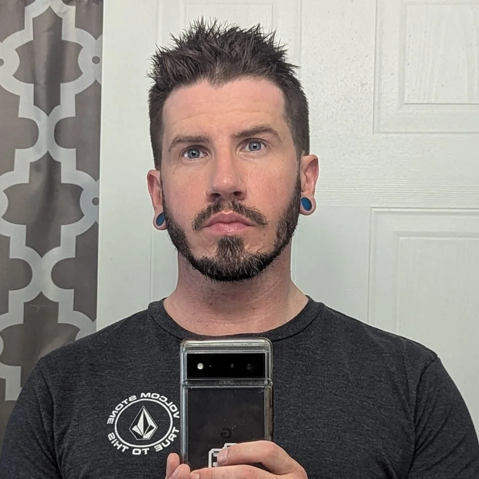 A man taking a mirror selfie indoors, wearing a dark t-shirt with a logo and sporting piercings and a trimmed beard.