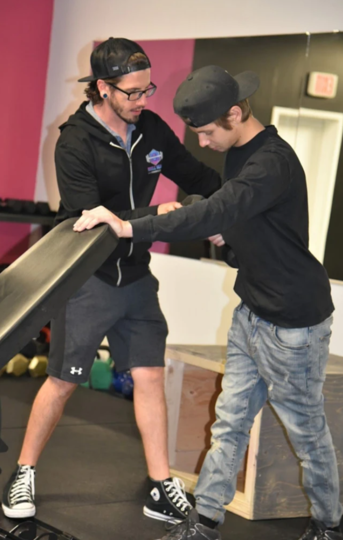 Two young men are practicing a self-defense move in a gym. One man is helping the other to practice an arm hold or choke hold on a padded training dummy. Both are wearing casual athletic clothing, including caps, and are focused on the training exercise.