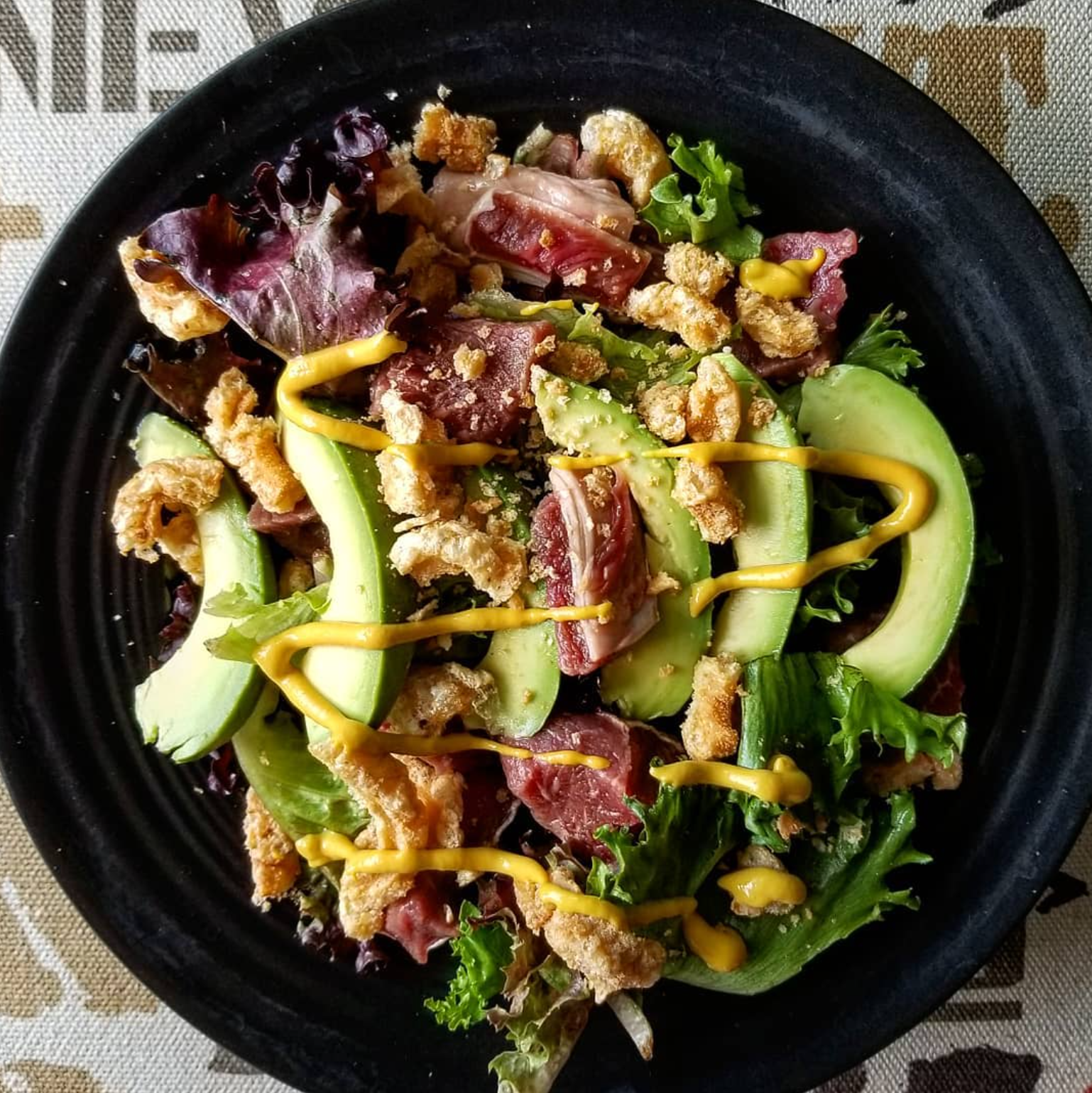 Mixed greens salad with avocado slices, crispy fried chicken, bacon, shredded cheese, and yellow mustard drizzle on black bowl.