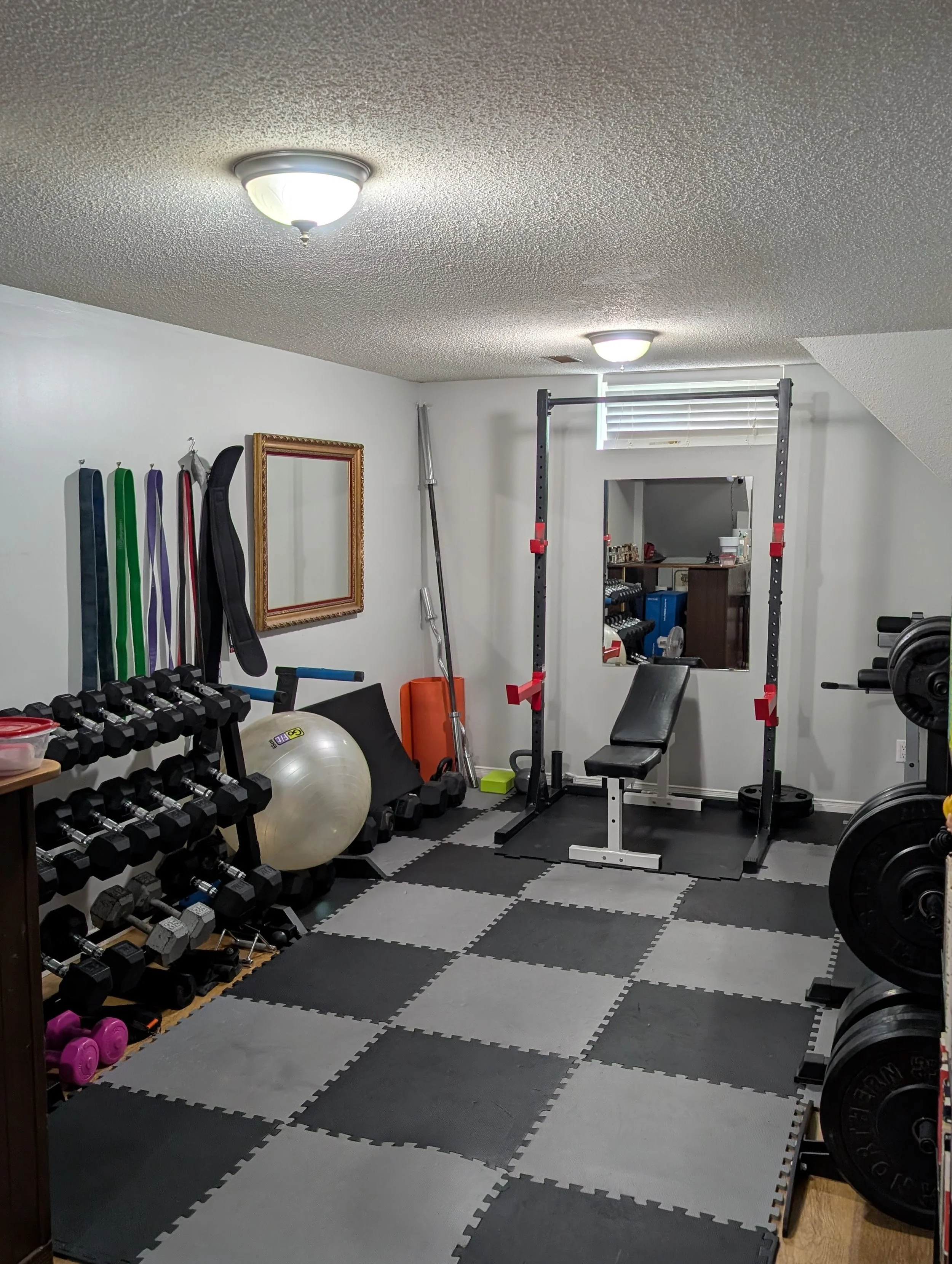 Home gym with black and white interlocking foam mats, various dumbbells, pink, purple, green, and blue resistance bands, a large exercise ball, a mirror, a weightlifting bench, a squat rack, and other exercise equipment.