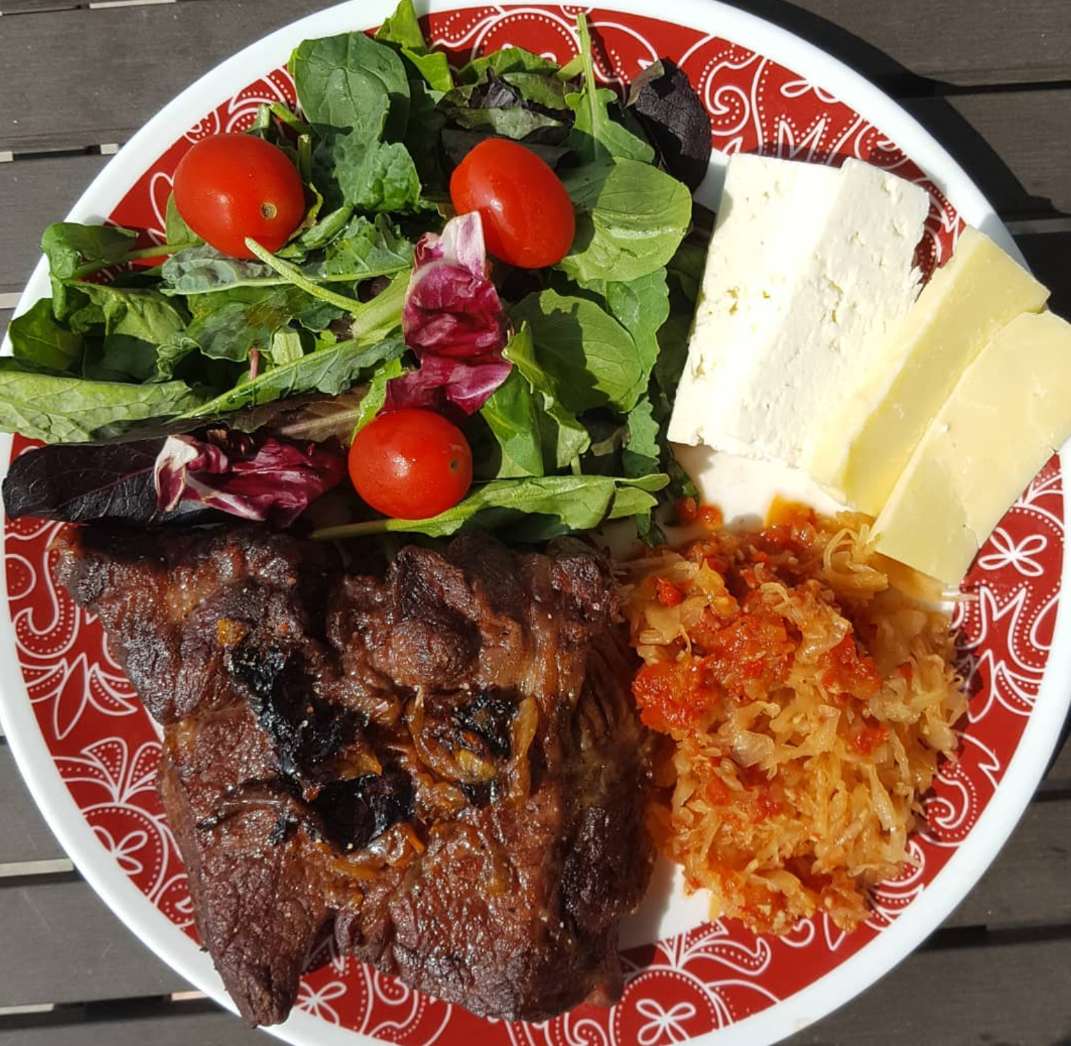 A festive plate with grilled steak, fresh green salad with cherry tomatoes, slices of cheese, and cooked cabbage with tomato sauce, all served on a decorative red and white plate.