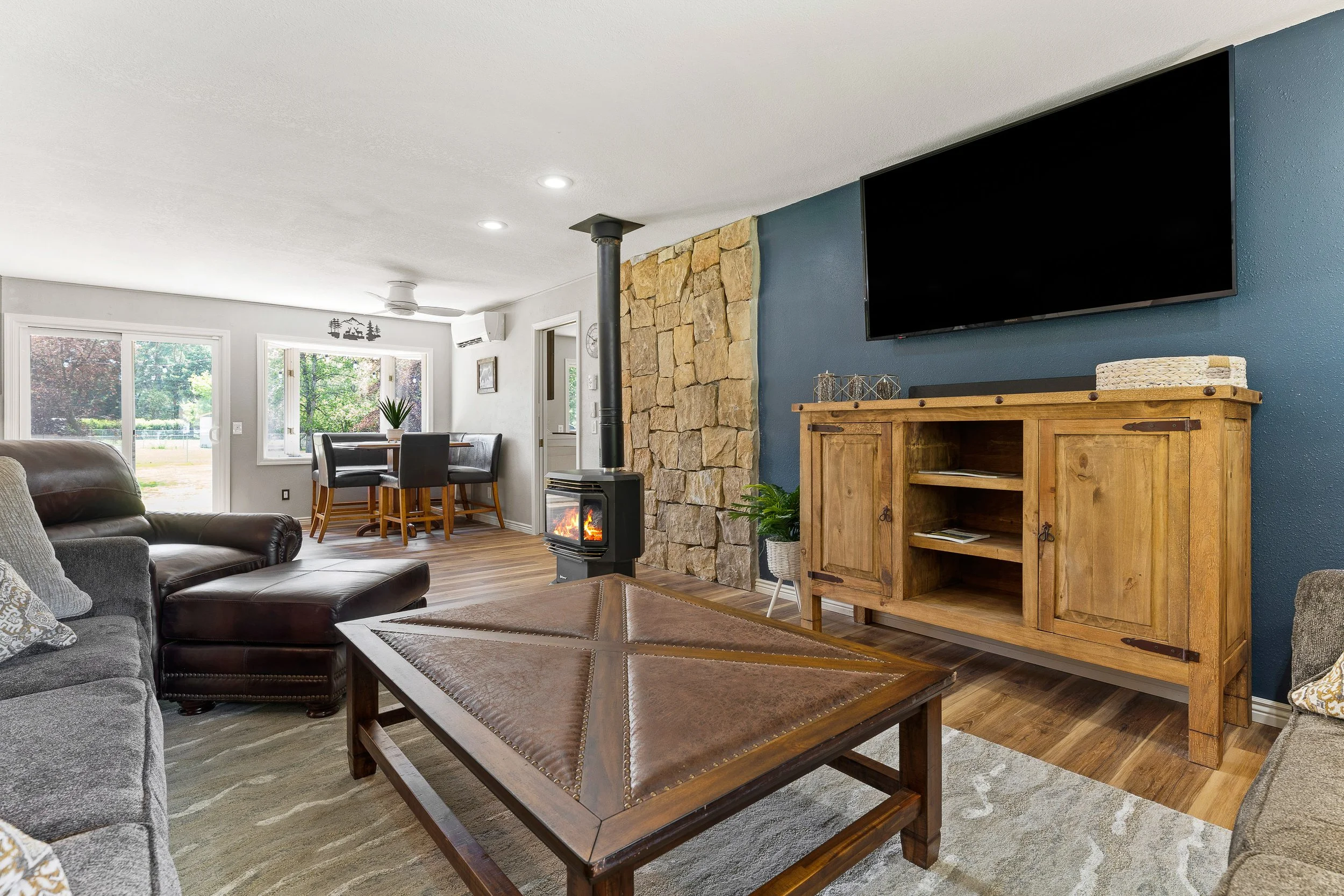 Living room with a leather sofa, wooden coffee table, TV mounted on a blue wall, stone accent wall, electric stove, dining table with chairs near windows, and various decorative items.