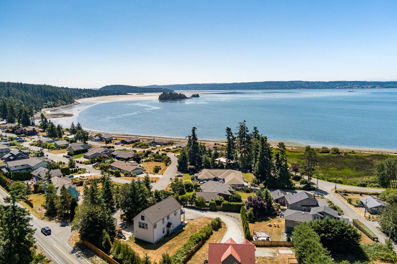 A coastal residential neighborhood with houses, trees, and streets, overlooking a calm bay with a beach and forested shoreline under a clear blue sky.