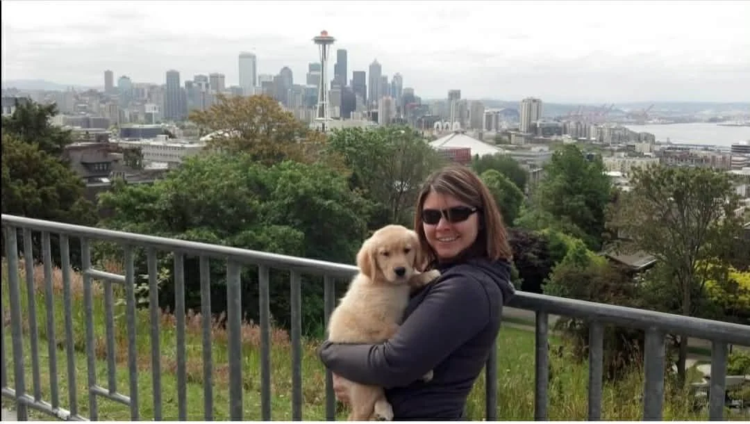 A woman with sunglasses holding a golden retriever puppy, with the Seattle skyline including the Space Needle in the background.