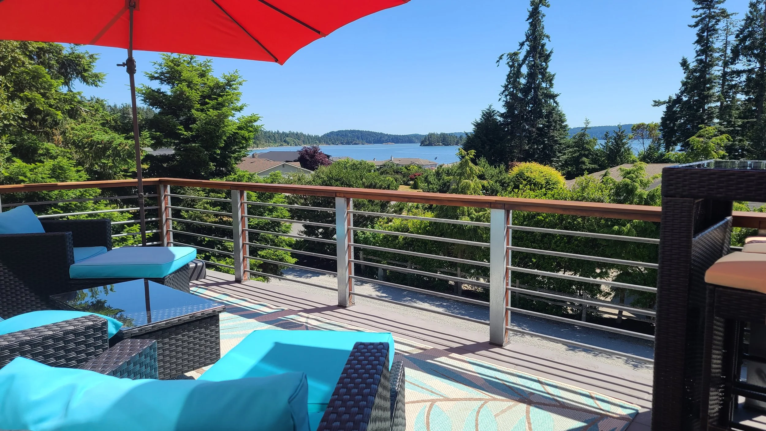 View of a balcony with outdoor furniture, including chairs with blue cushions and a glass-top table, overlooking lush green trees, a body of water, and distant hills under a clear blue sky.
