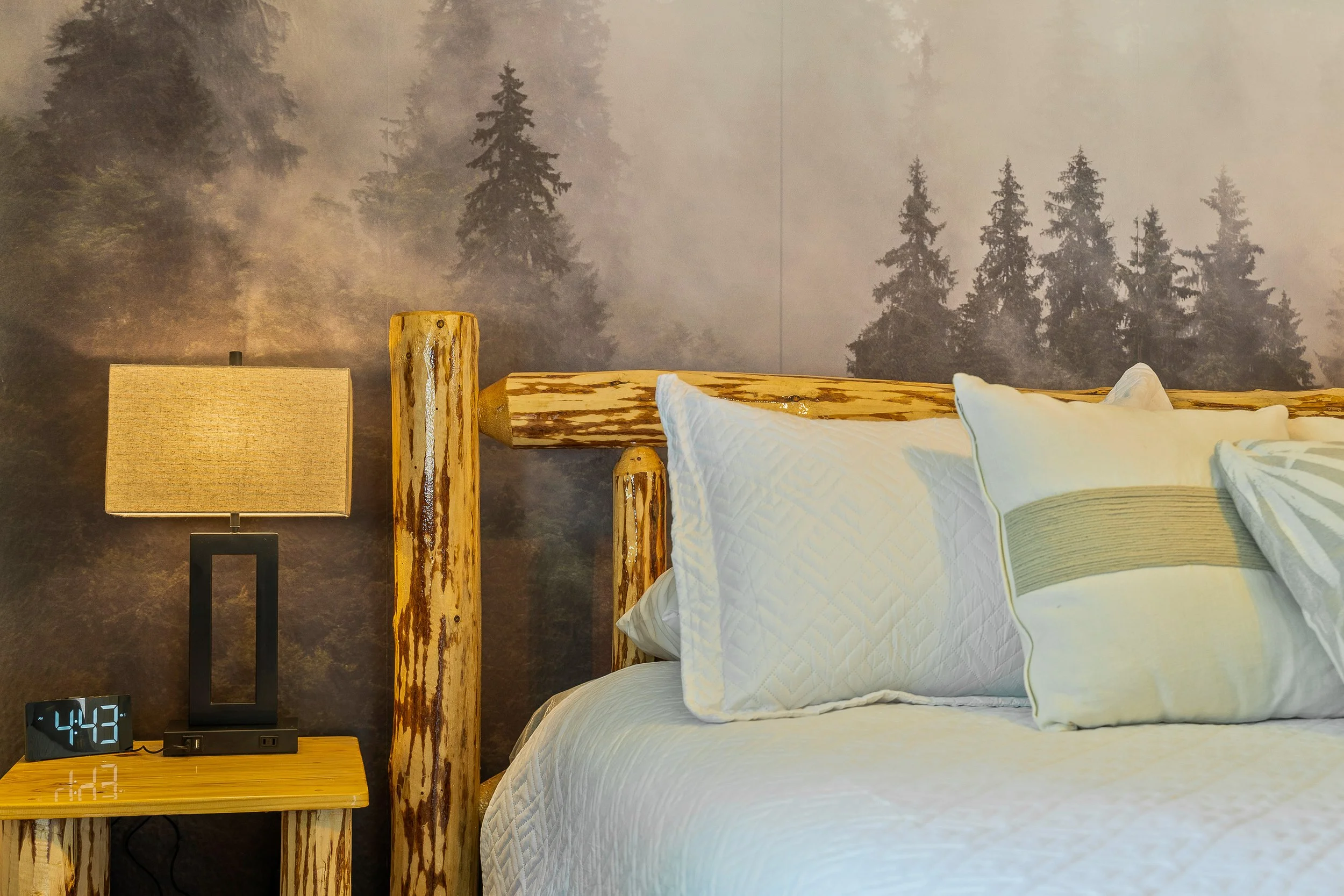 A bedroom with a forest-themed wallpaper, displaying trees in a misty forest. The bed has white bedding with multiple pillows. To the left, a nightstand holds a digital clock showing 4:43 and a modern table lamp, with wooden logs used as decor.
