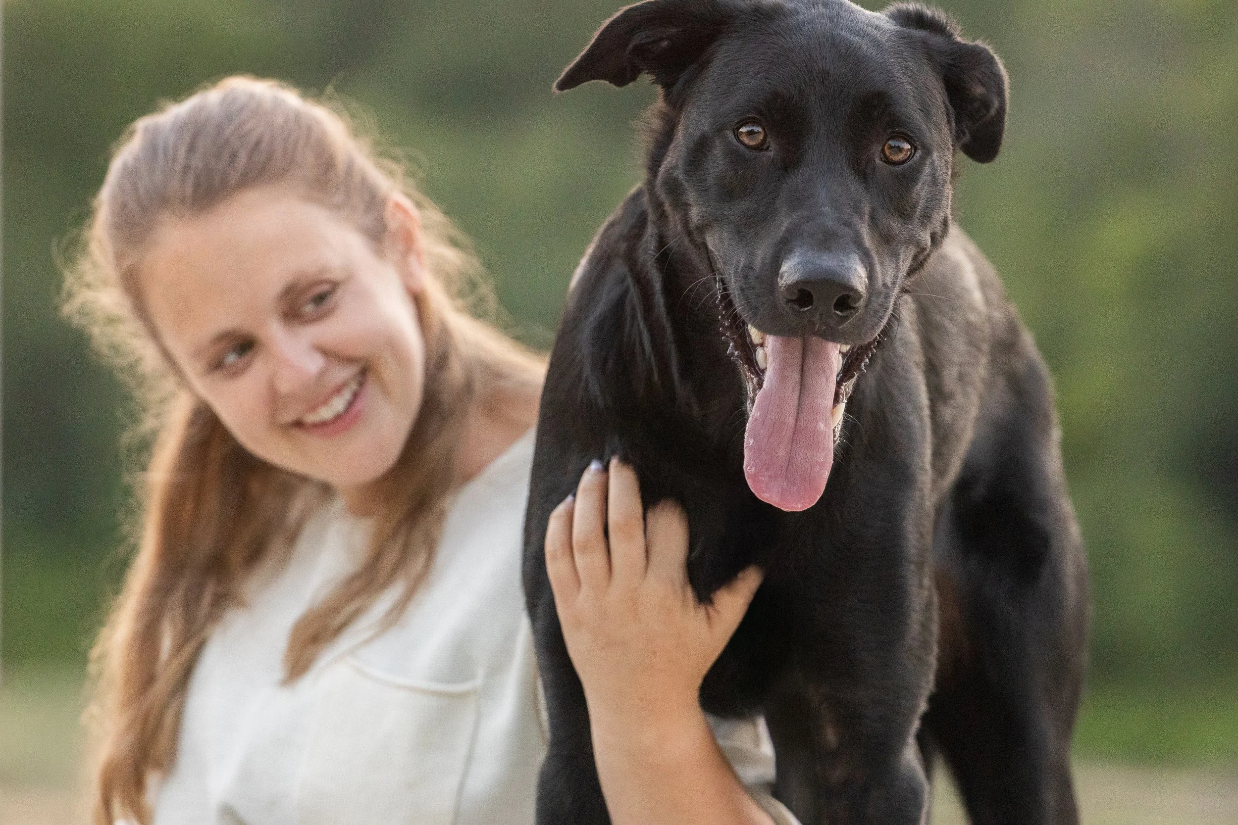 A woman with long hair and a white top holding and smiling at a black dog with its tongue out, outdoors with a blurred green background.