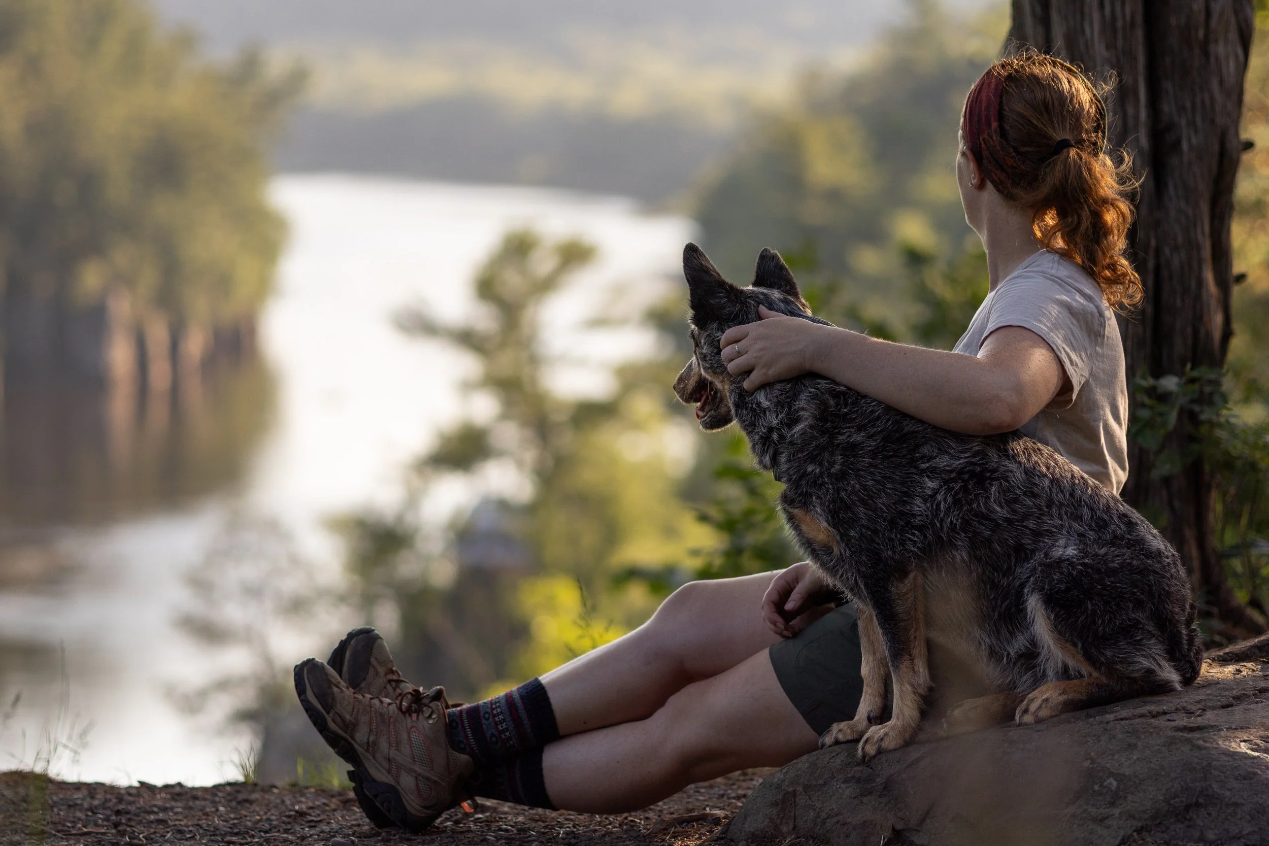 A woman with red hair tied back, sitting by a riverbank with her legs extended and holding a dog on her lap. She is wearing hiking shoes and shorts, and the scene is serene with trees and water in the background, illuminated by soft natural light.
