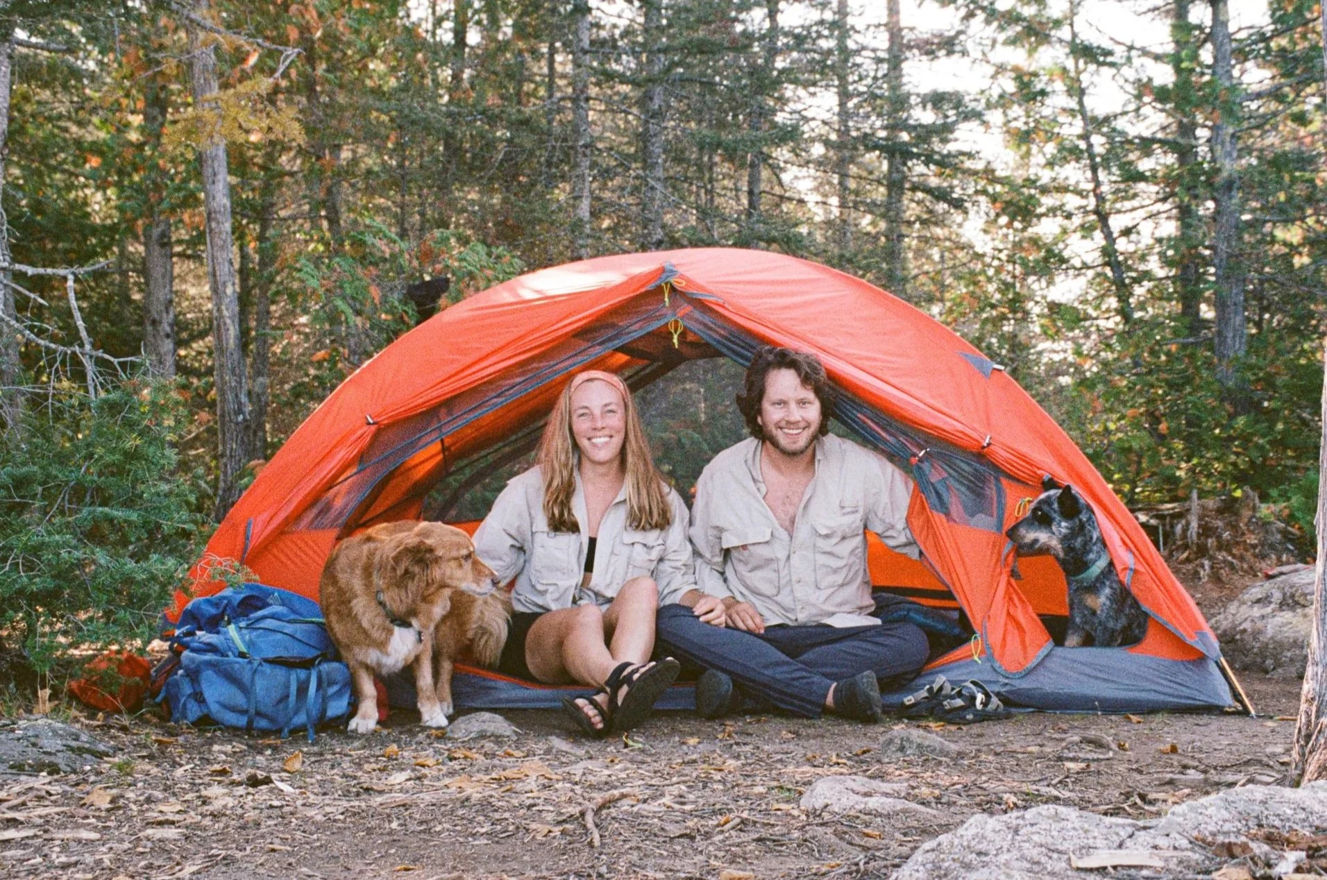 Two people sitting inside an orange camping tent in a forest, surrounded by trees. They are smiling, with dogs beside them, and camping gear is visible around the tent.