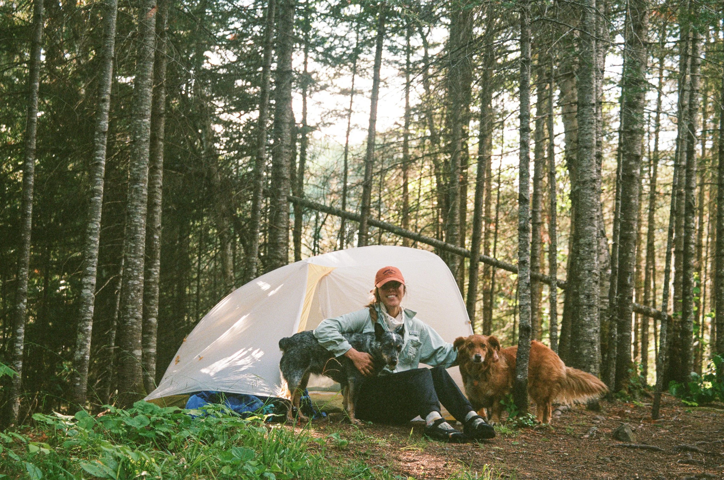 A woman sitting outside a tent in a forest, smiling, with a dog beside her. The woman is wearing glasses, a hat, and outdoor clothing. It appears to be daytime with sunlight filtering through trees.