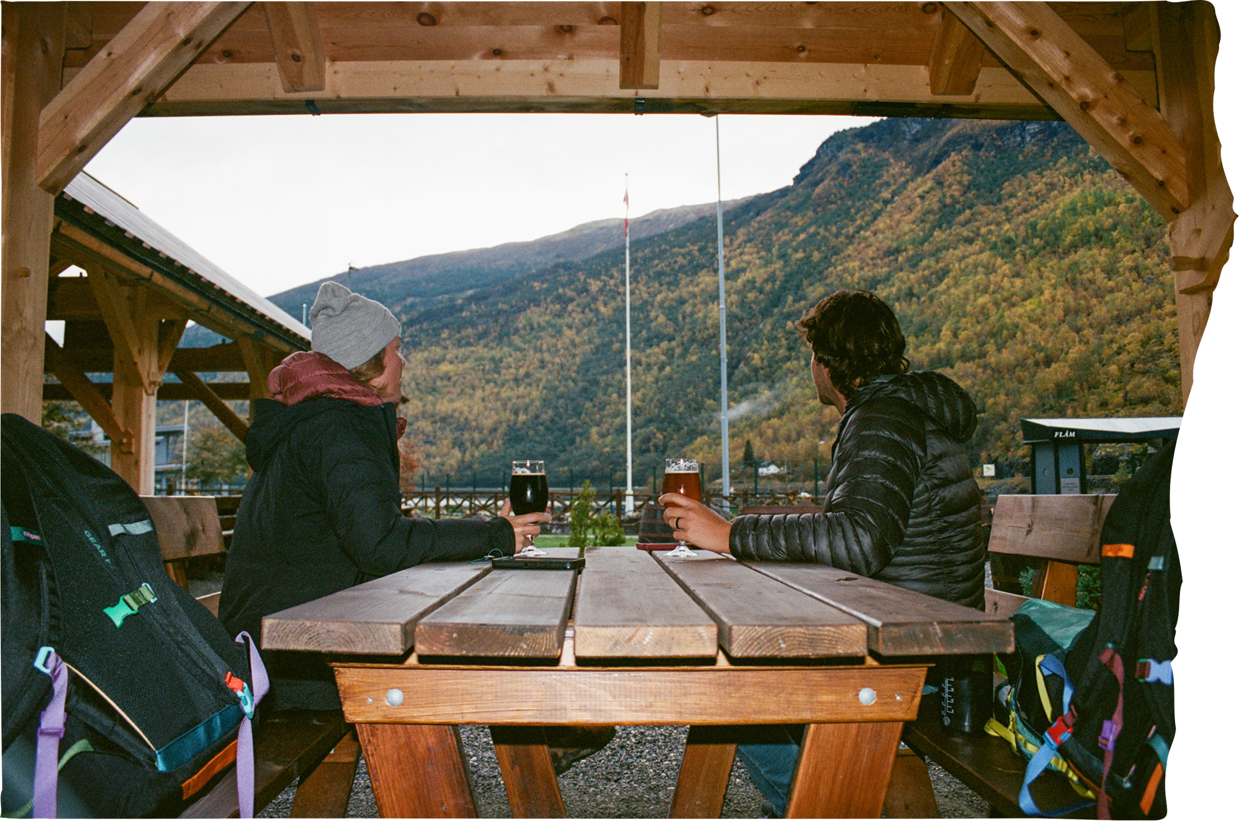 Two people sitting at a wooden table outdoors, drinking beers, with a mountain and autumn foliage in the background.