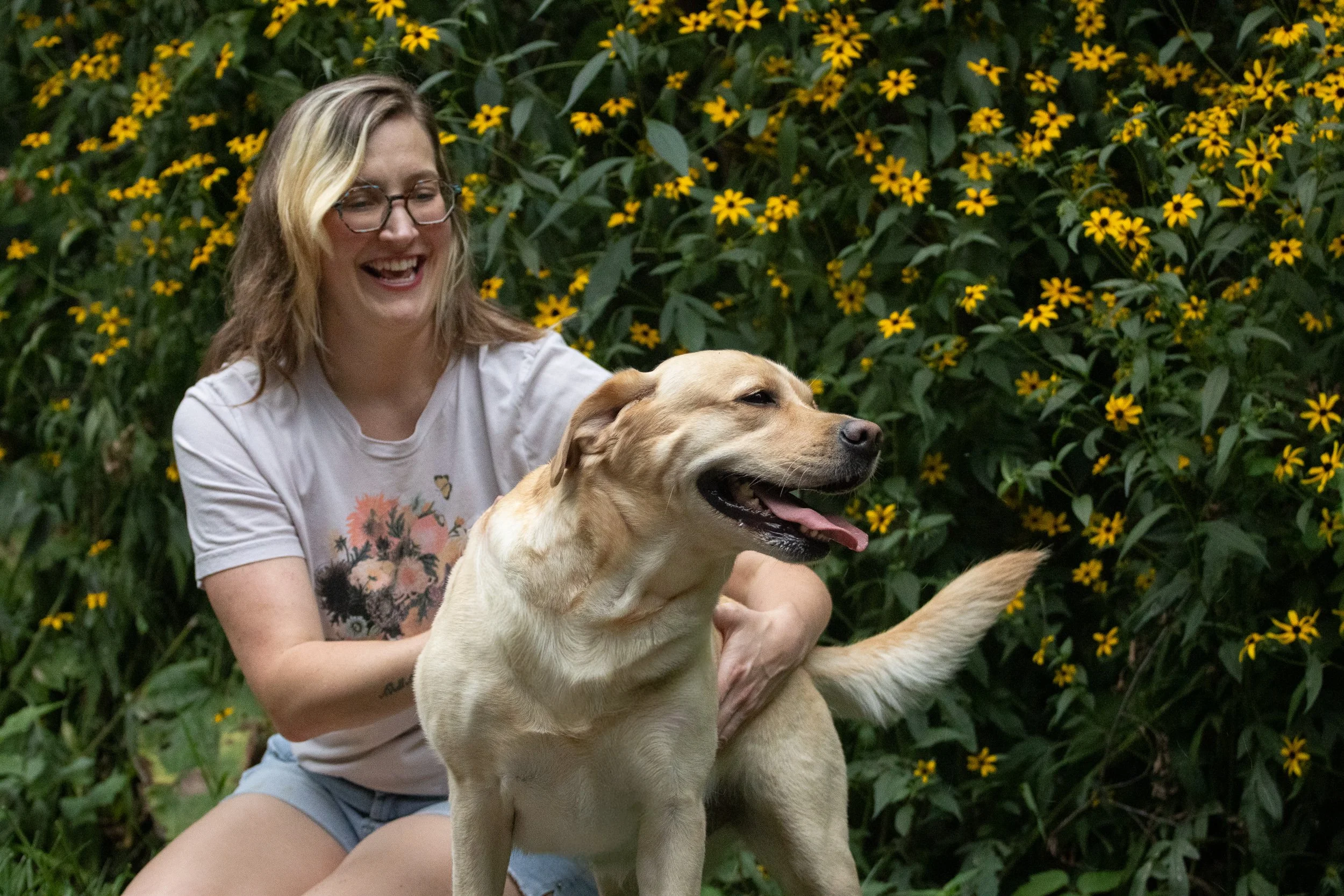 A woman with glasses and blonde hair smiling while hugging a large yellow Labrador Retriever dog outdoors in front of yellow flowers.