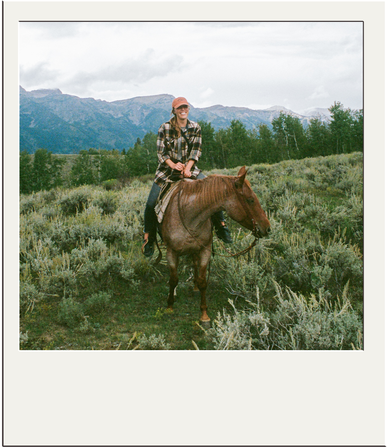 A woman riding a brown horse through a grassy field with mountains and trees in the background, wearing a plaid shirt and a baseball cap.