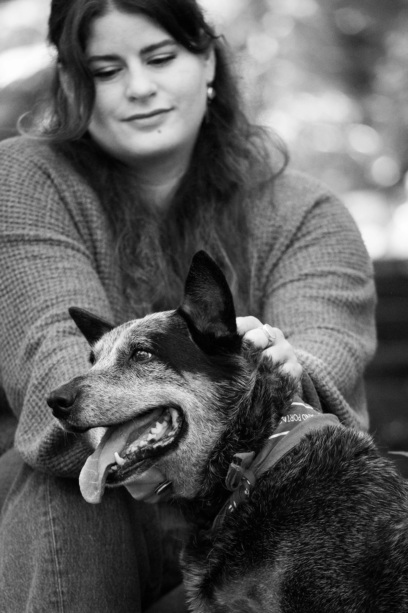 A woman with long dark hair and earrings smiling while petting a large dog with a thick coat, a pattern on its collar, and an open mouth showing a happy expression. The photo is in black and white.