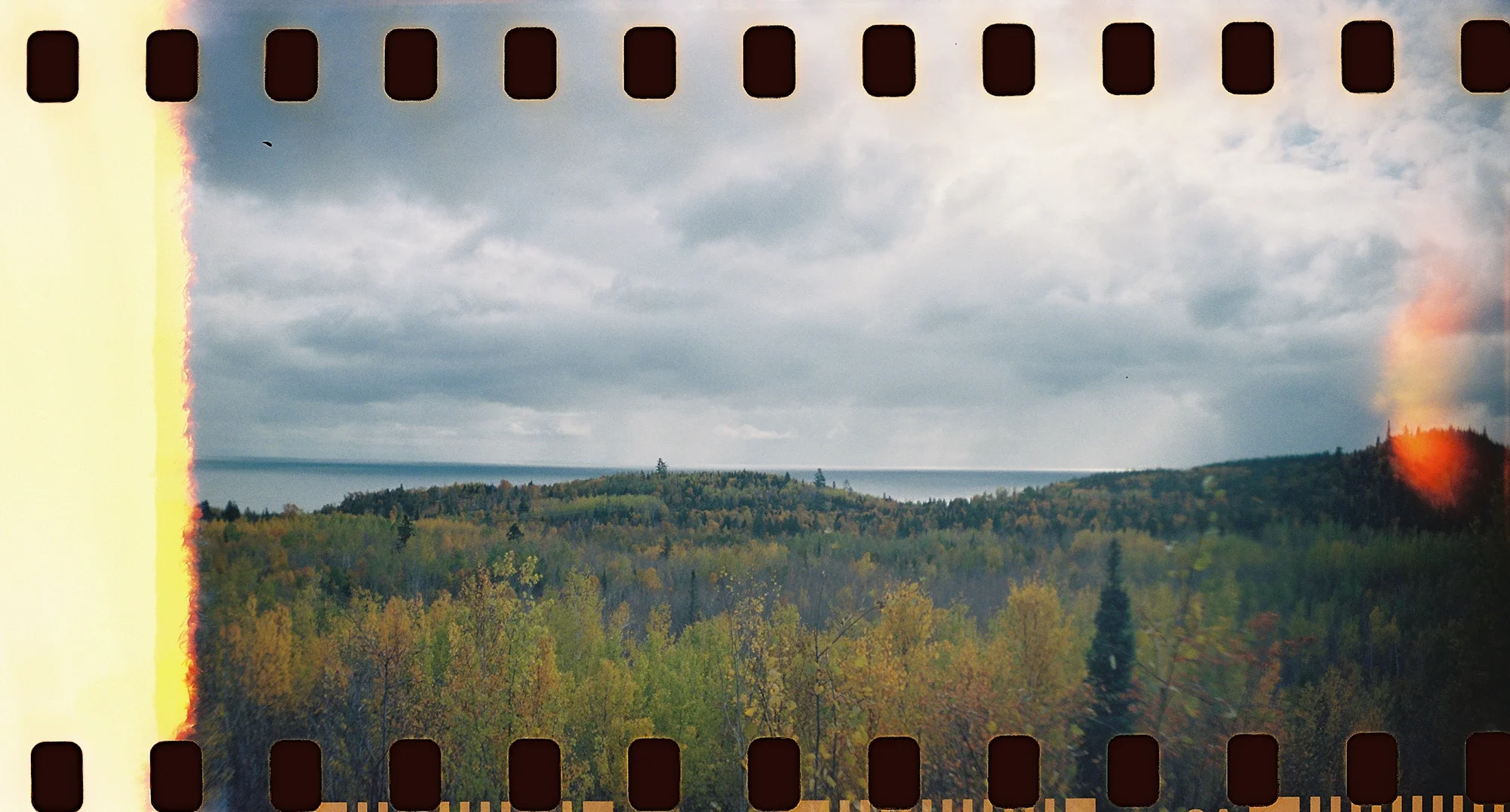 A scenic view of a forested landscape with a body of water in the distance under a cloudy sky, and a faint rainbow on the right side.