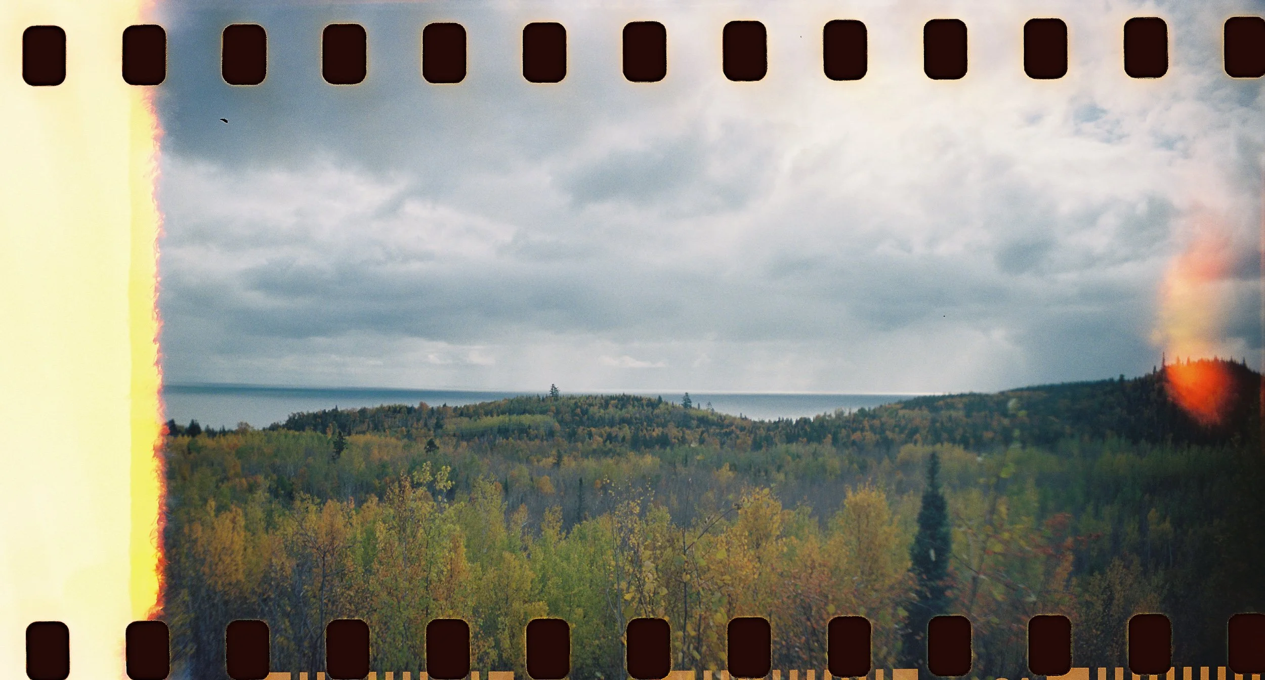 A landscape with a cloudy sky over a forested hillside, and a body of water in the distance. The photo has visible film light leak on the left and right edges.