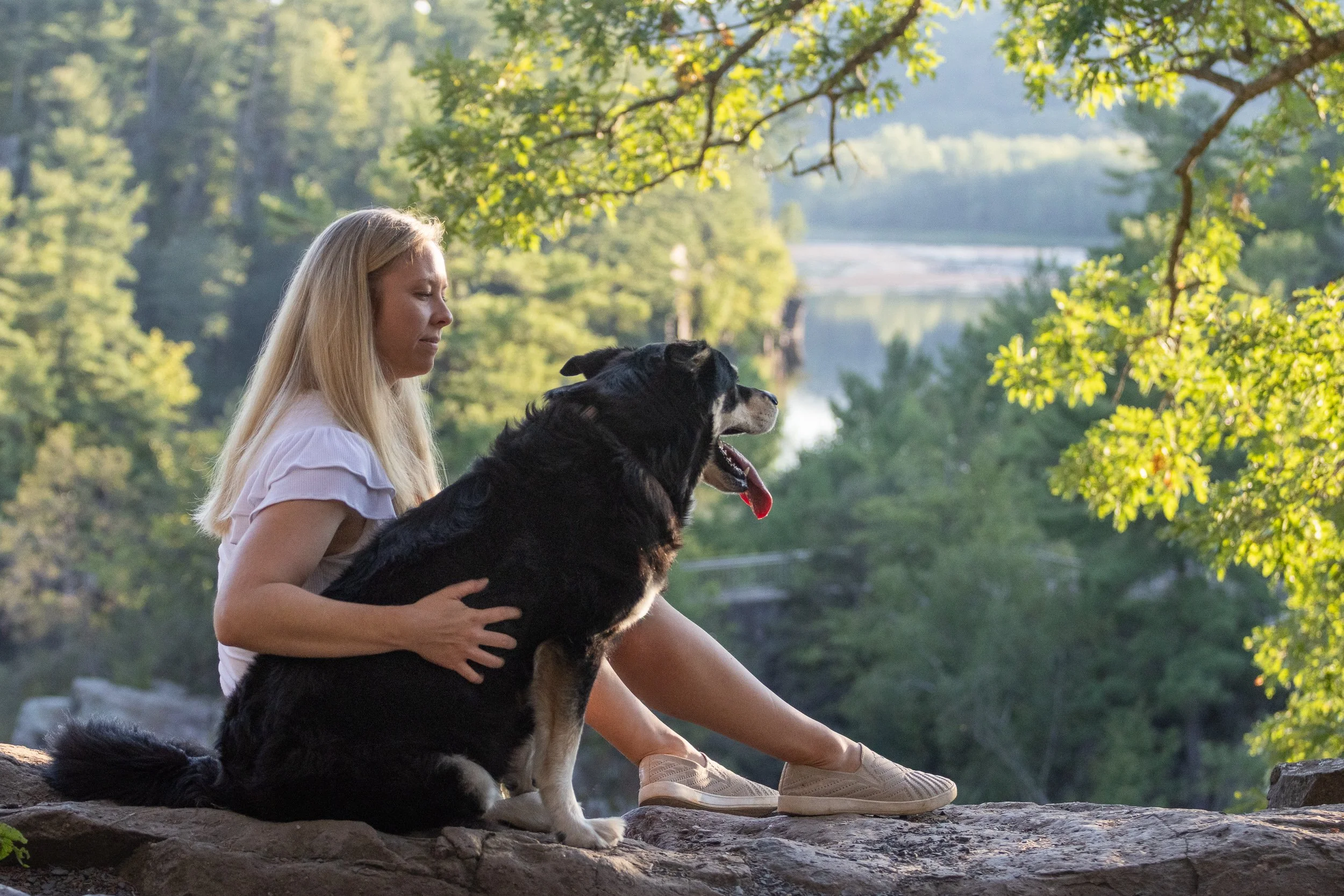 A woman with long blonde hair sitting outdoors on rocks, holding a black and tan dog, with a river and green trees in the background.