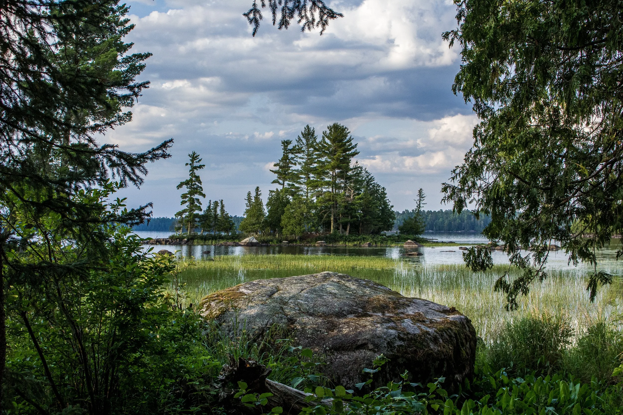 A scenic view of a lake in the Boundary Waters of northern Minnesota with trees and rocks in the foreground and a cloudy sky overhead.