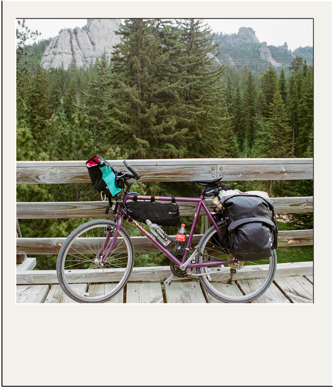 A purple bicycle with cargo bags on the front and back, parked against a wooden fence on a bridge overlooking a forested mountain landscape.