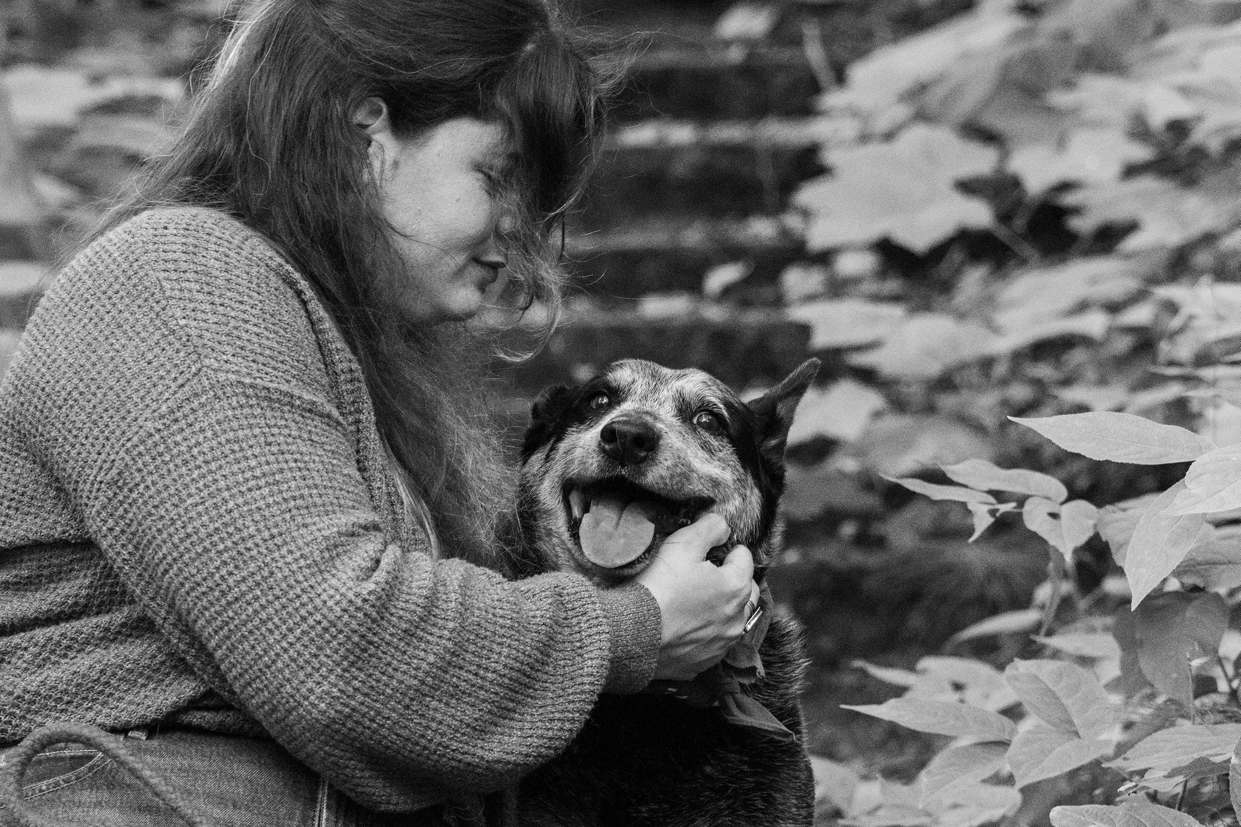 A woman with glasses and long hair holding a smiling dog with its tongue out in an outdoor setting with trees and foliage.