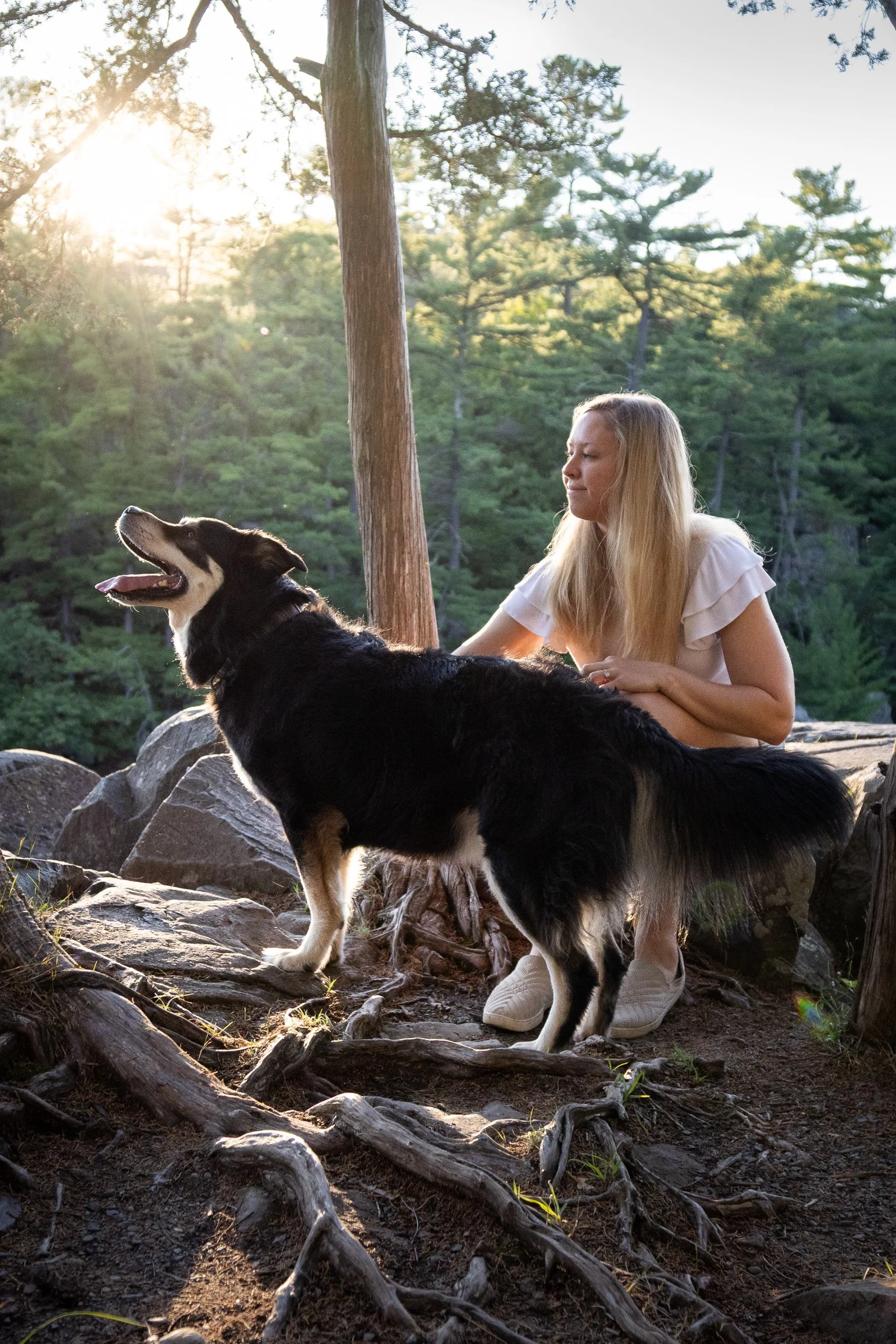A woman with long blonde hair sitting outdoors on rocks near a tree, smiling at her black and tan dog, with a forest in the background and sunlight filtering through the trees.