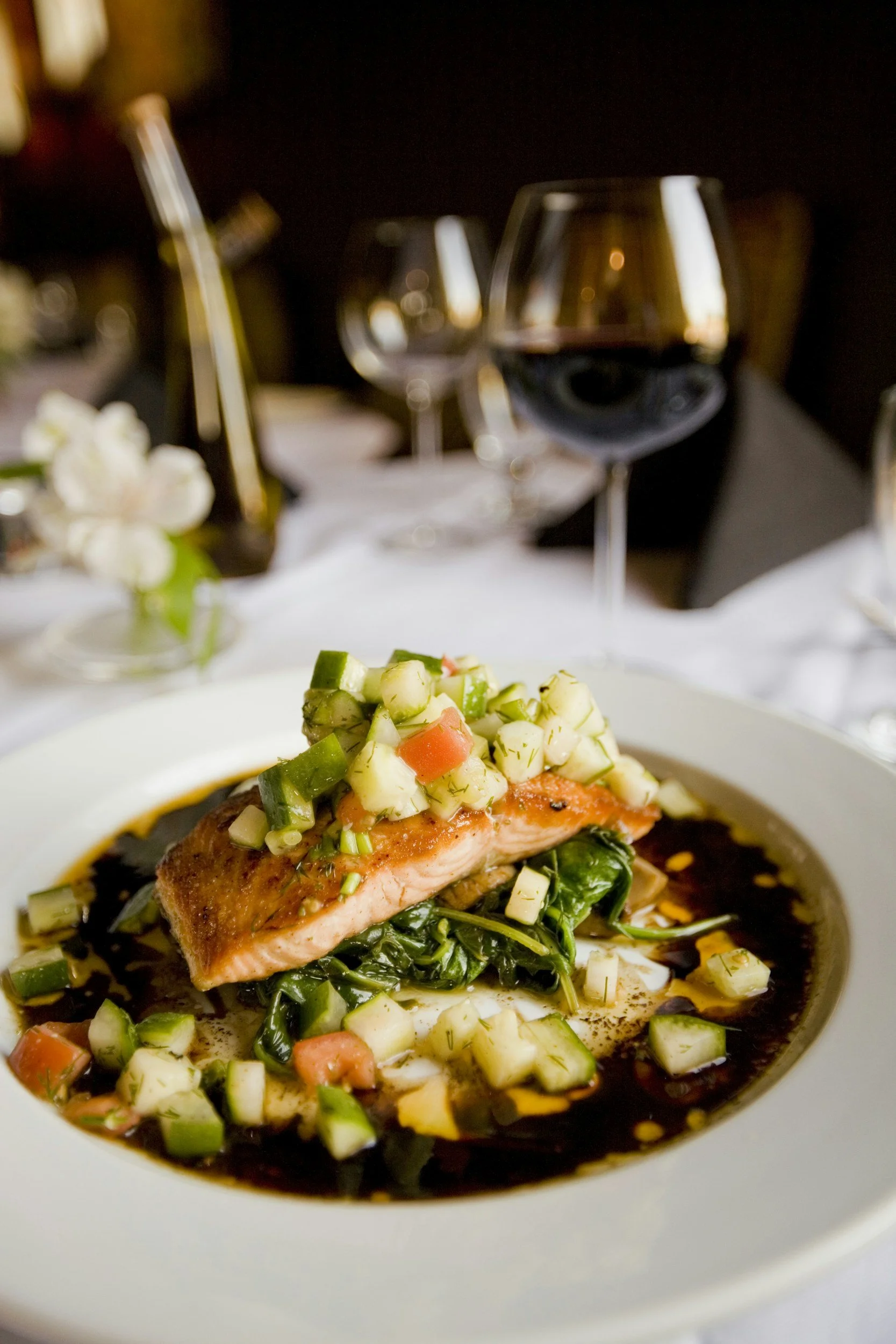 Close-up of a plated dish with cooked salmon, diced vegetables, and sauce, on a white plate. In the background, two glasses of red wine and a dark, elegant dining setting.