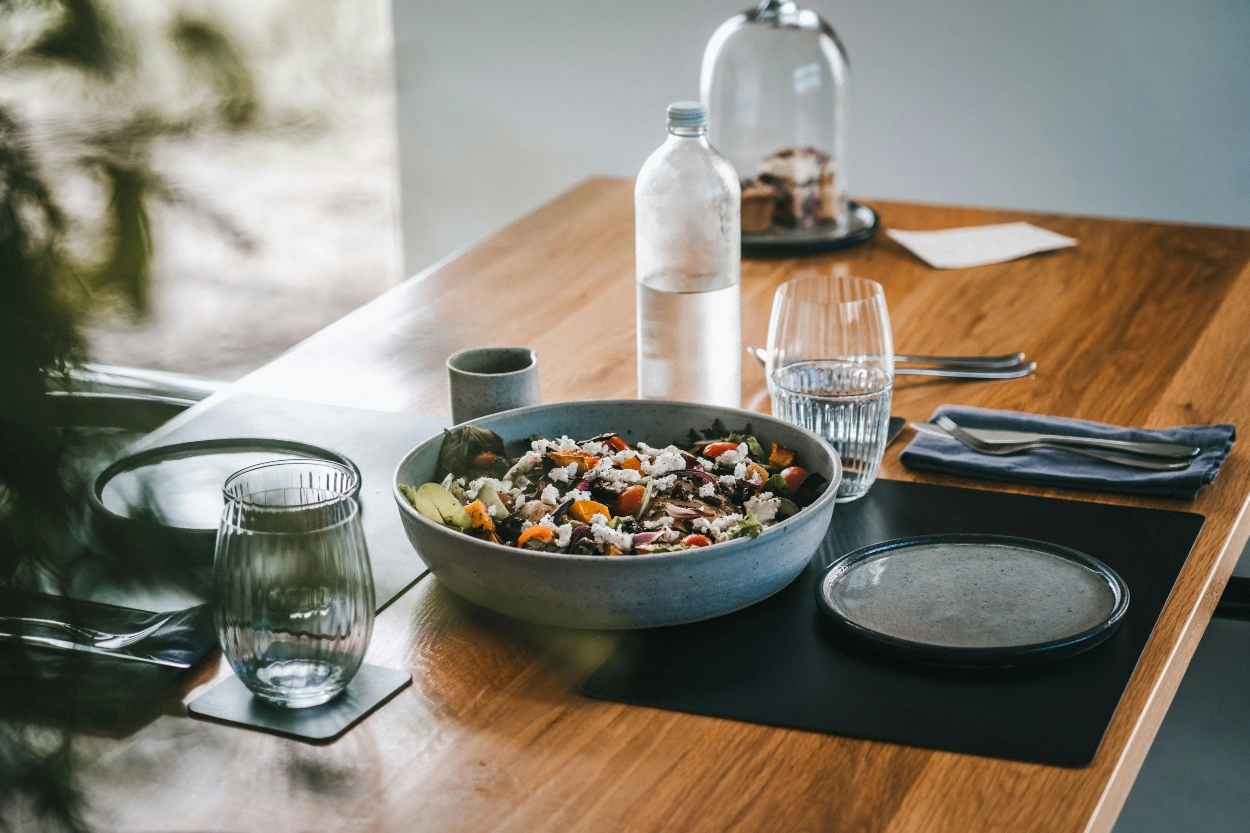 A wooden dining table set with a large bowl of colorful salad, two glasses of water, a gray napkin with silverware, a clear water bottle, and a glass dome with cookies inside. A piece of paper is on the table near the wall.