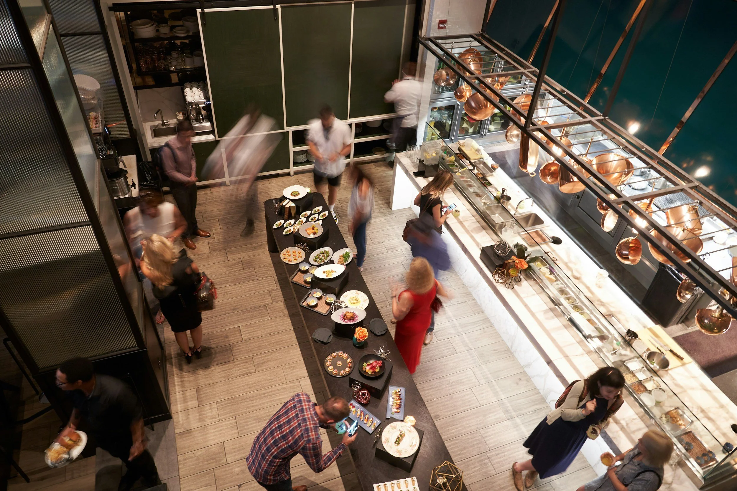 A top-down view of a busy buffet-style restaurant with customers and staff. There are tables with plates of food, a counter with various dishes, and kitchen staff preparing meals. Copper hanging pots and blue wall decor are visible.