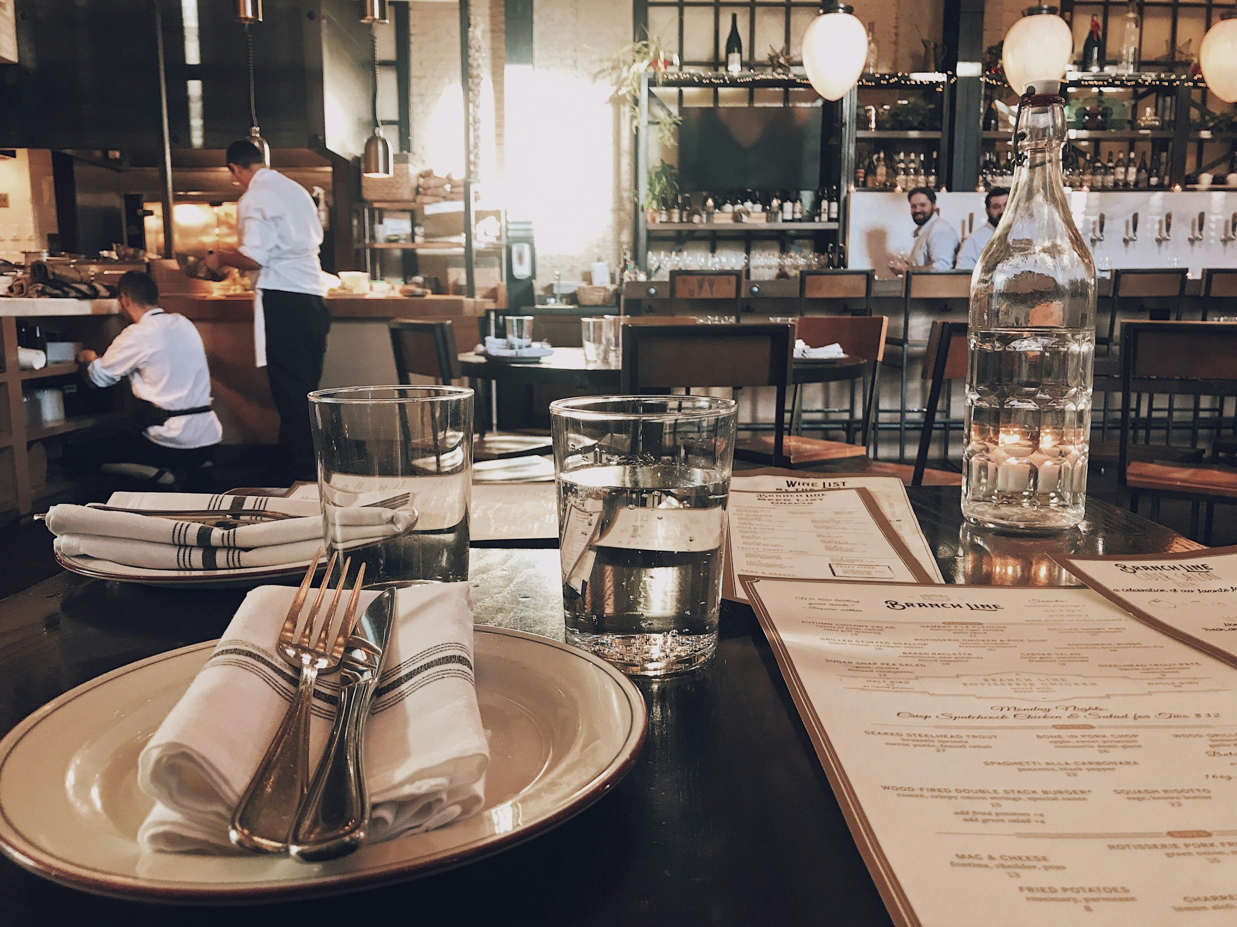 Inside a restaurant with a table setting including glasses of water, cutlery, napkins, and a menu, with chefs working in the background in a modern, stylish dining area.