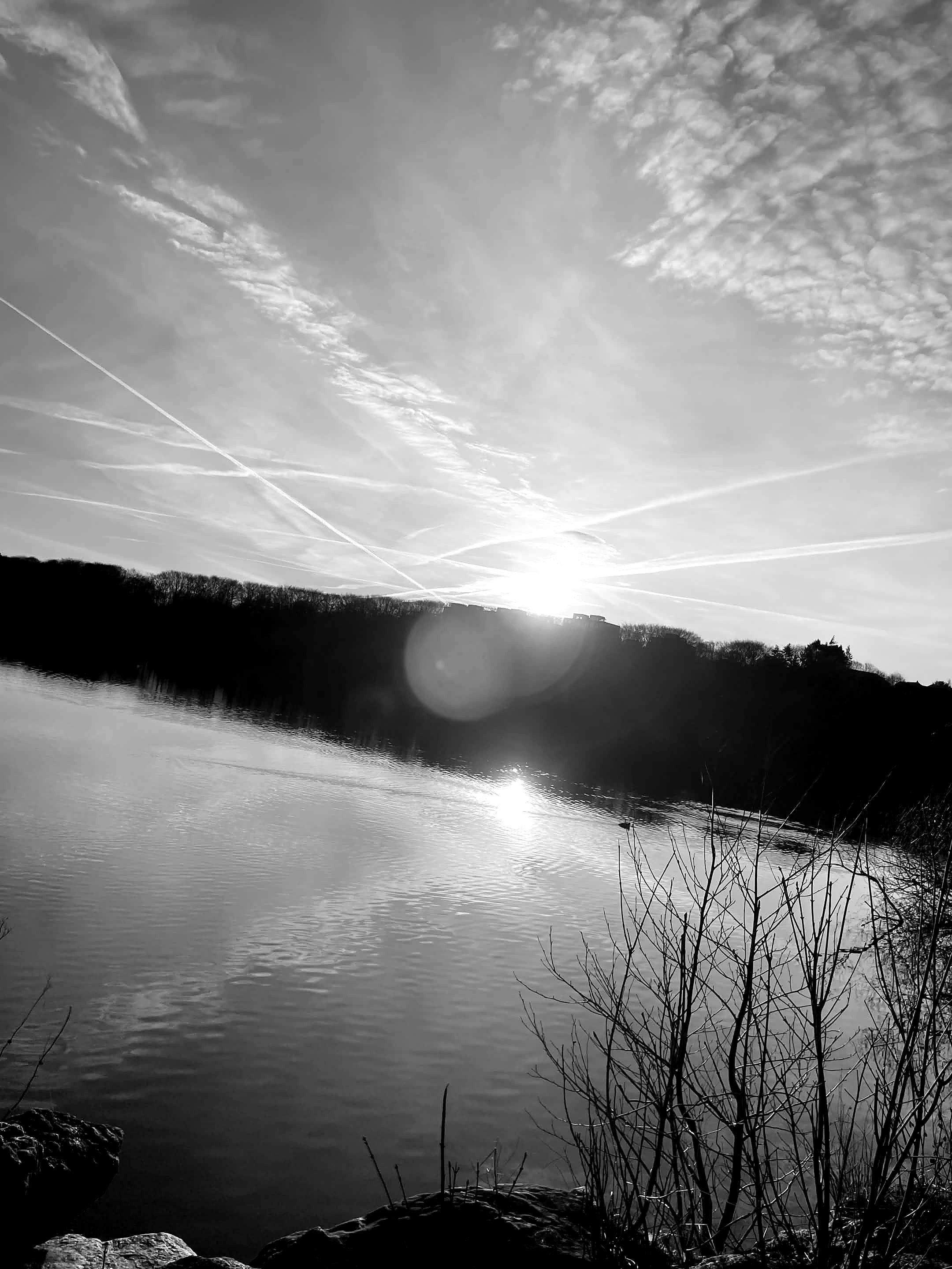 A black and white photo of a river at sunset with trees on the horizon and a sky filled with clouds and contrails.