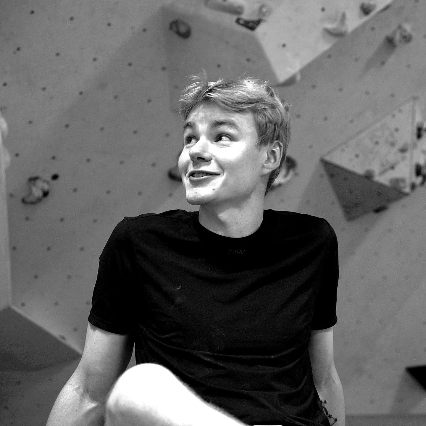 A young man with short, tousled hair wearing a black t-shirt, smiling and looking to his left, sitting in front of a climbing wall in an indoor gym.