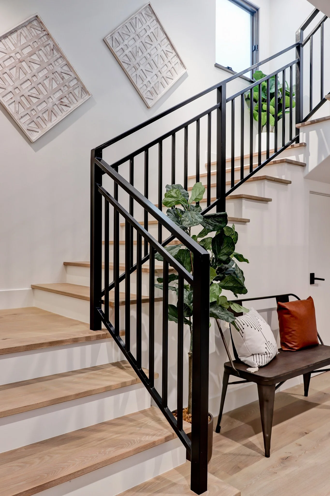 Modern staircase with light wooden steps, black metal railing, and white walls. Decor includes framed wooden wall art, a tall potted plant with large green leaves, and a seating area with pillows.