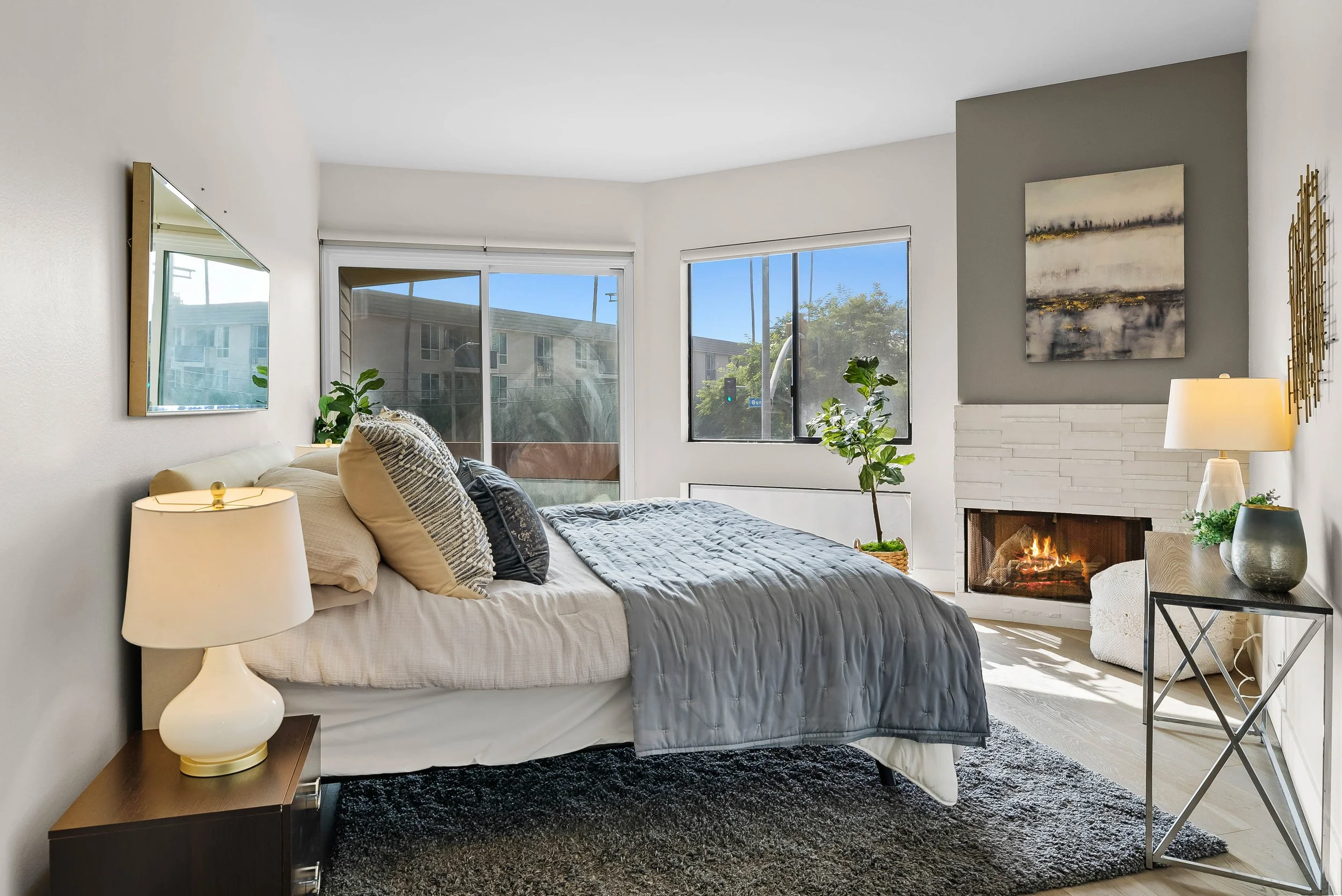 A modern living room with a bed, nightstand, fireplace, and large windows showing a sunny day outside.
