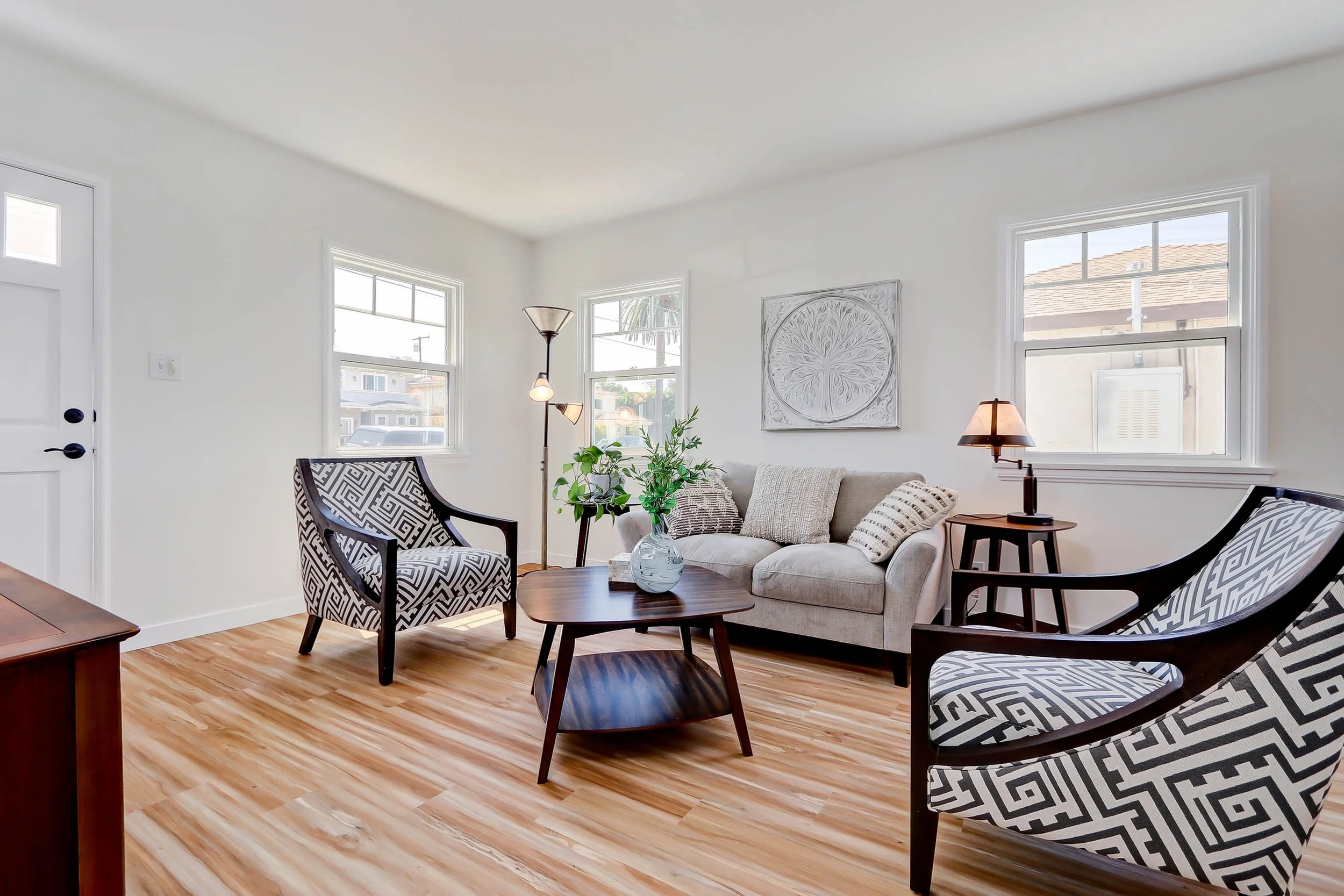 Living room with white walls and wooden floor, decorated with patterned armchairs, a beige sofa, a small round wooden coffee table with a glass vase, and various lamps. Large windows allow natural light inside, and there is a square wall art piece.
